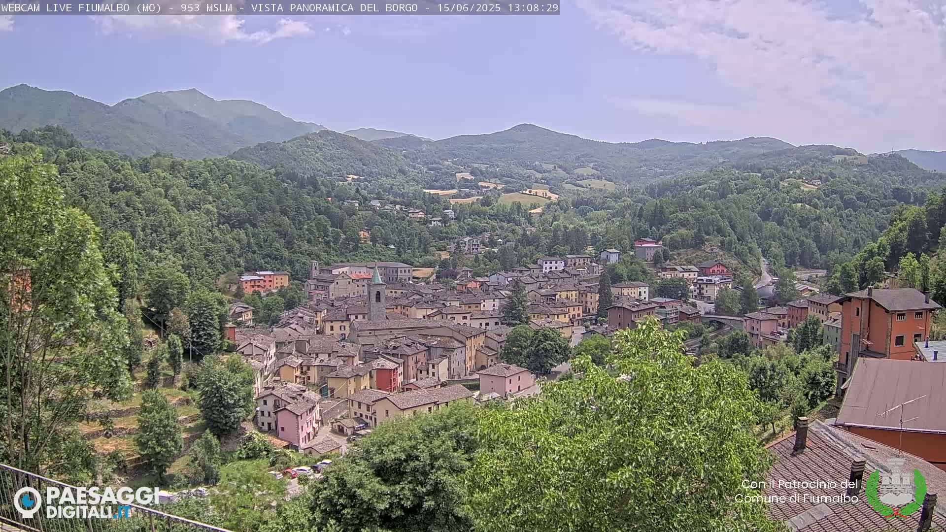 A quaint village nestled in a valley, surrounded by lush green hills and mountains under a partly cloudy sky.