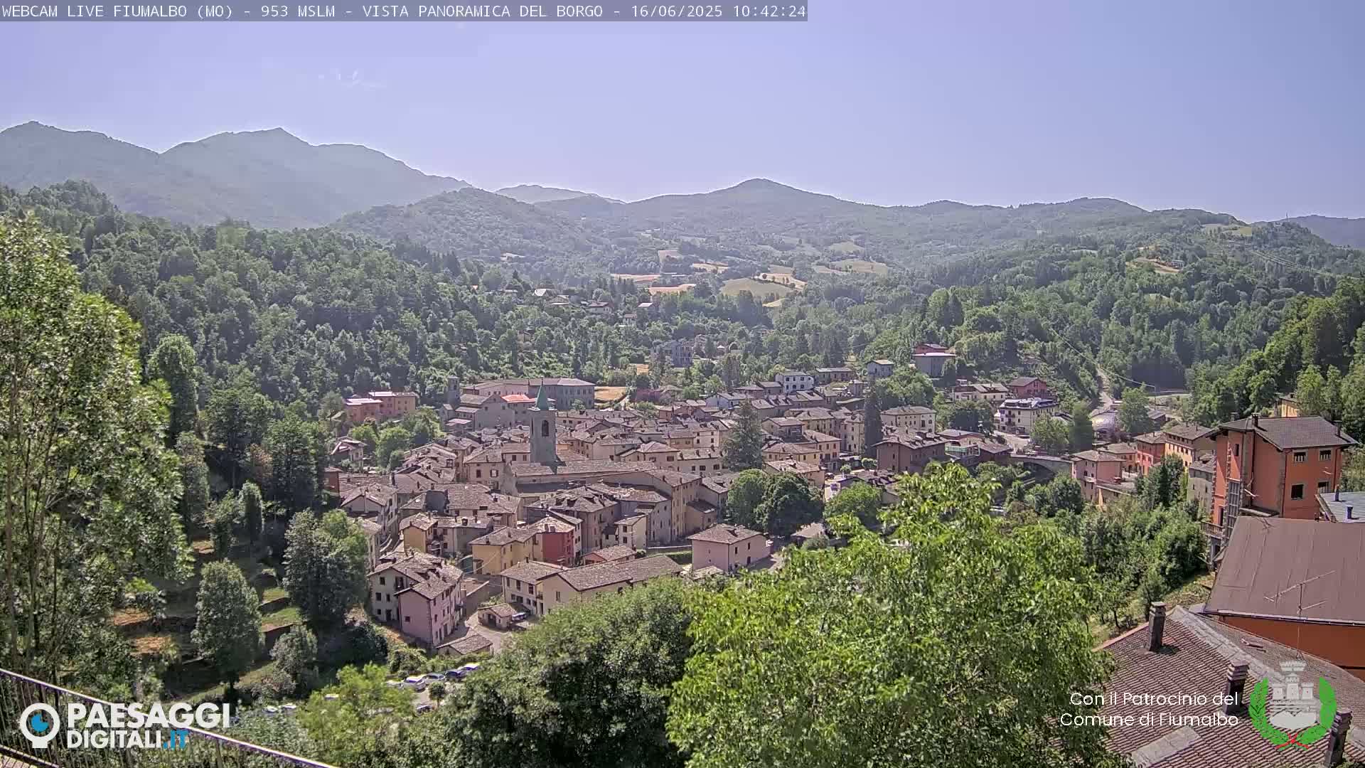 A panoramic view shows a small town nestled in a valley surrounded by lush green hills and mountains under a clear blue sky.