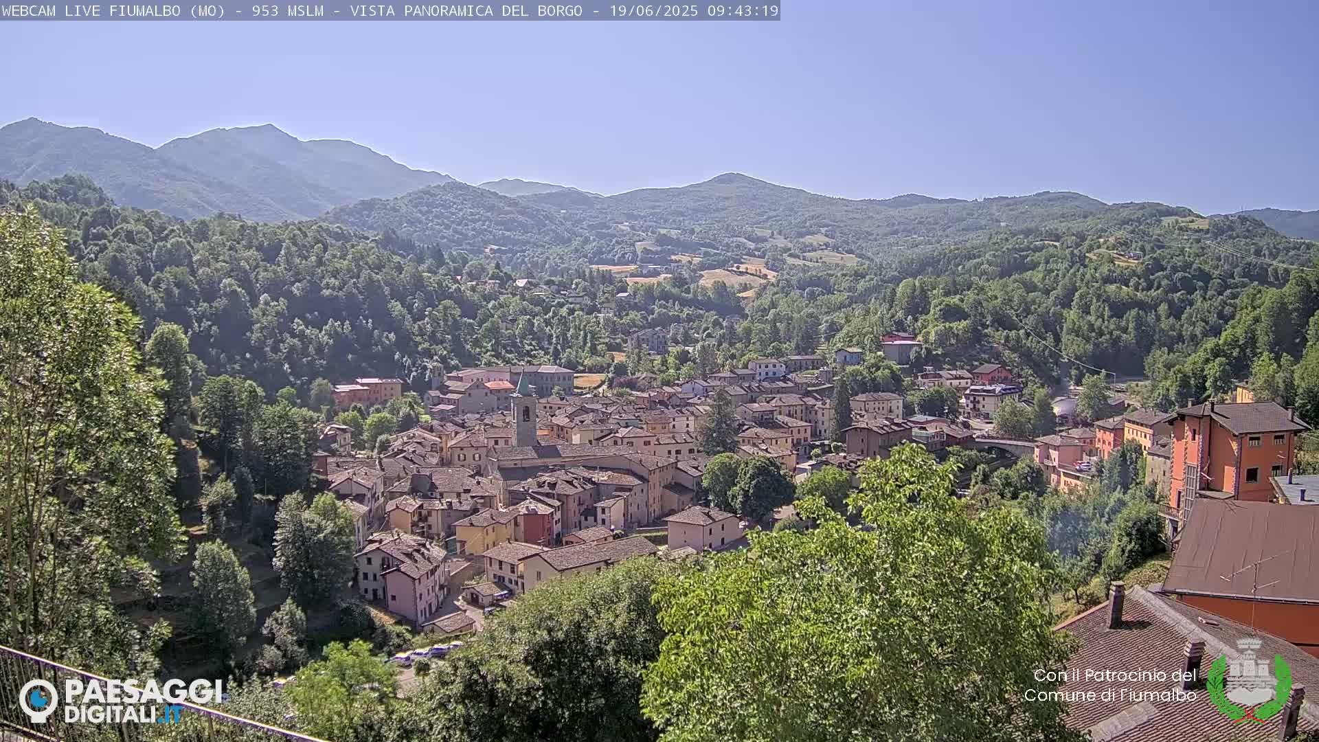 A panoramic view shows a town nestled in a valley surrounded by lush green hills and mountains under a clear, sunny sky.