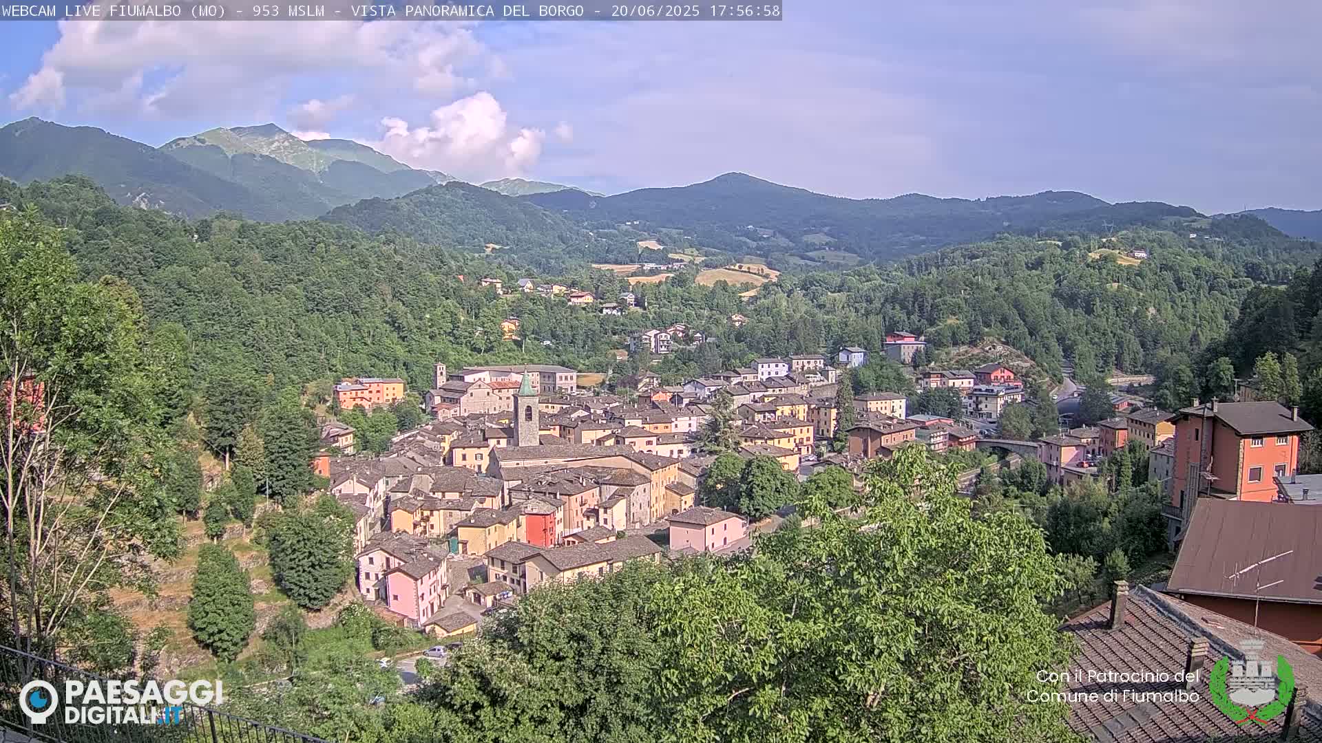 A partly cloudy day reveals a small town nestled in a valley surrounded by lush green hills and mountains.