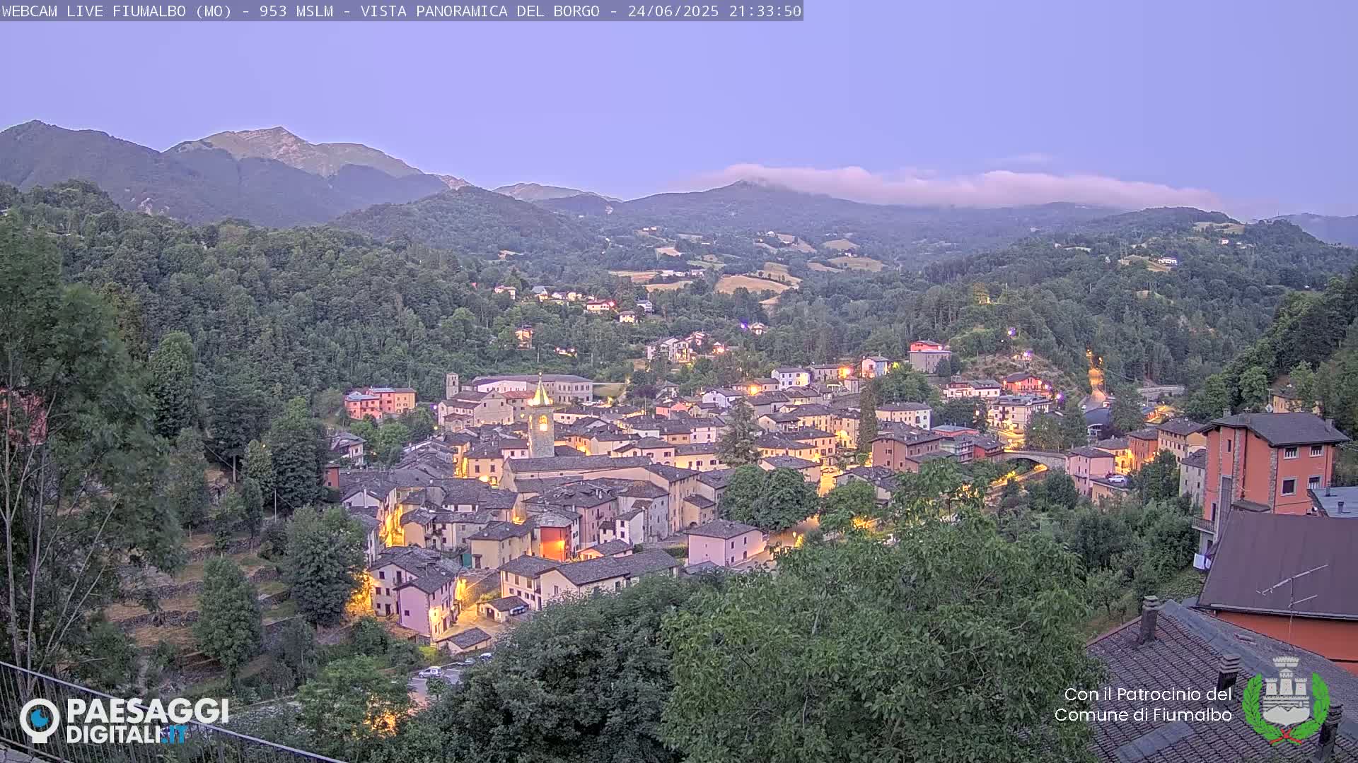A small, illuminated town nestled in a valley between forested hills under a twilight sky with some high clouds.
