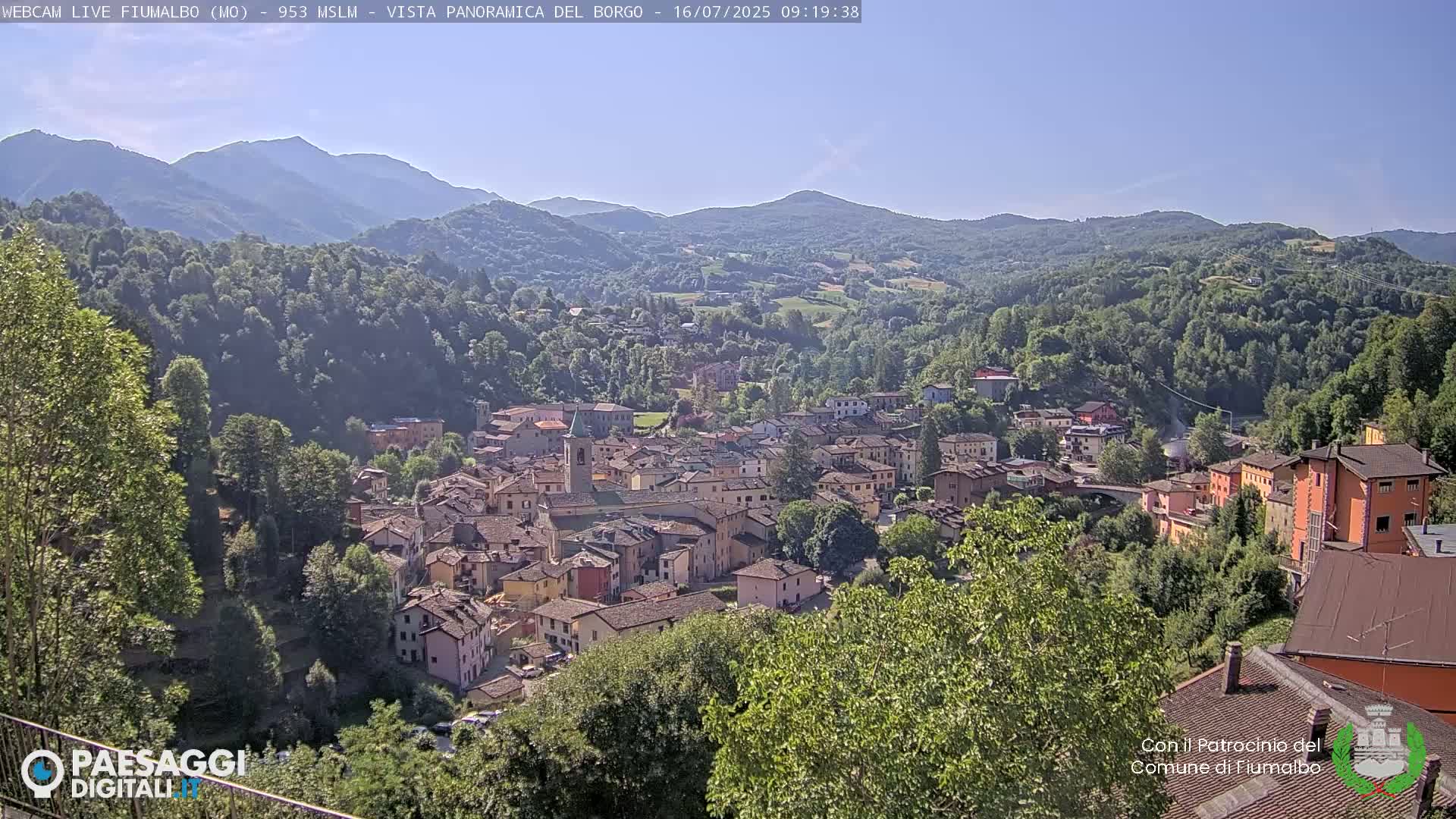 A panoramic view shows a village nestled in a valley surrounded by lush green hills and mountains under a clear blue sky.