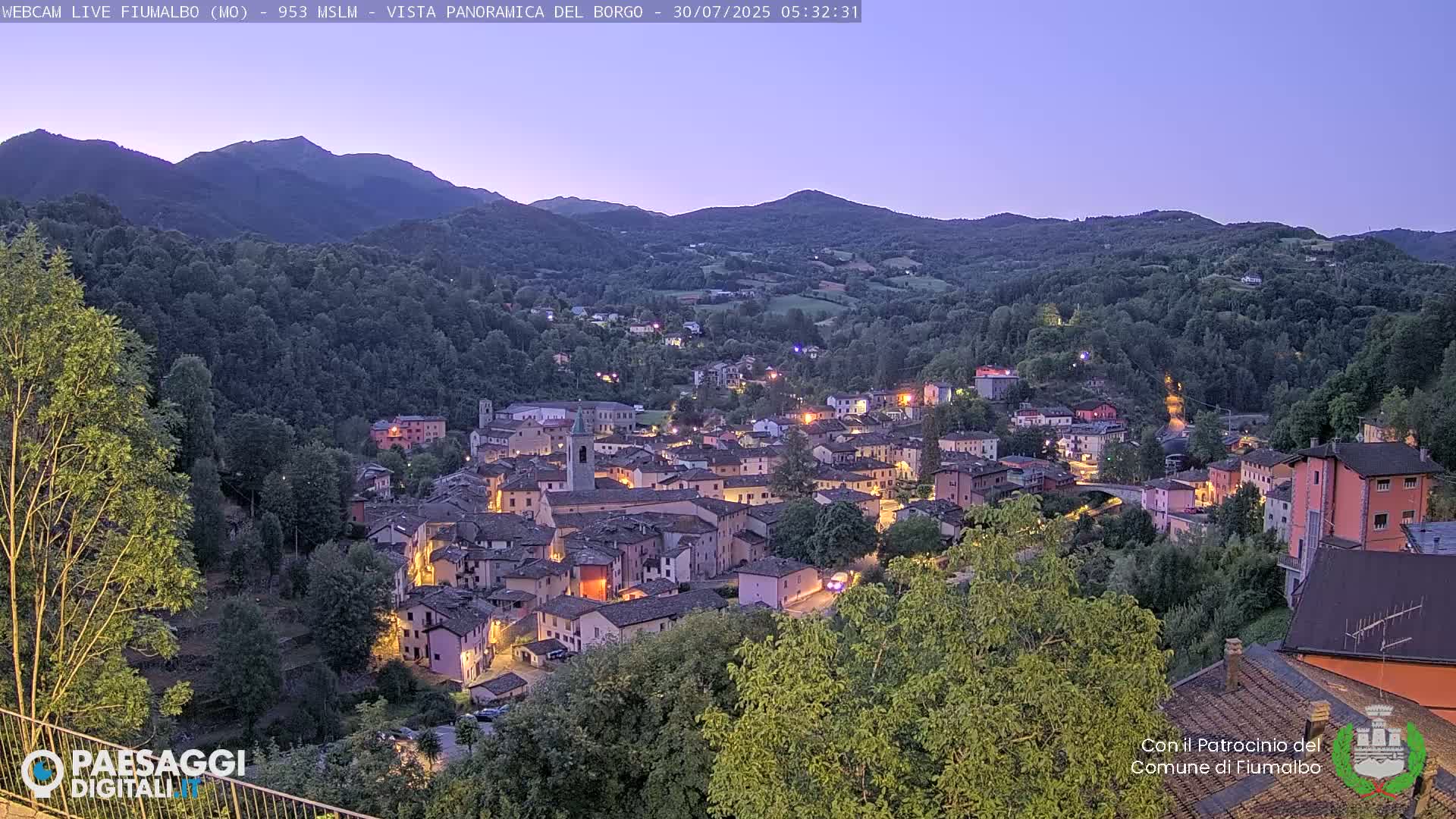 A picturesque village nestled in a valley surrounded by lush green hills and mountains under a partly cloudy sky.