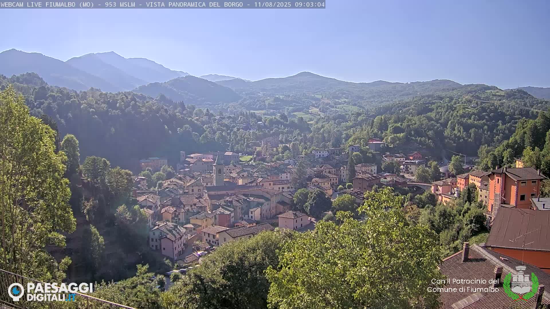 A panoramic view shows a town nestled in a valley surrounded by lush green hills and mountains under a clear blue sky.