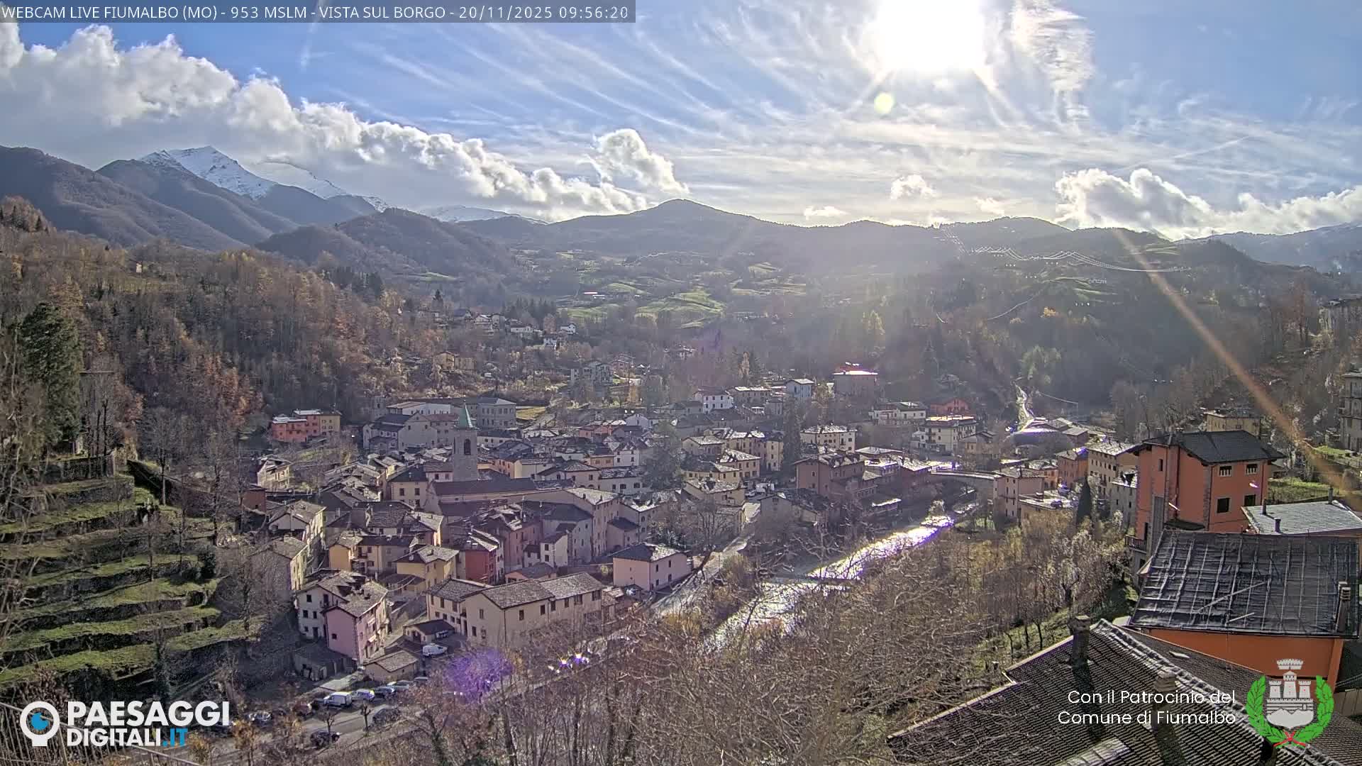 A vibrant mountain village, featuring colorful buildings and a winding river, is nestled among forested hills with distant snow-capped peaks, all bathed in bright sunshine under a partly cloudy blue sky, indicating clear and crisp late autumn or early winter weather.