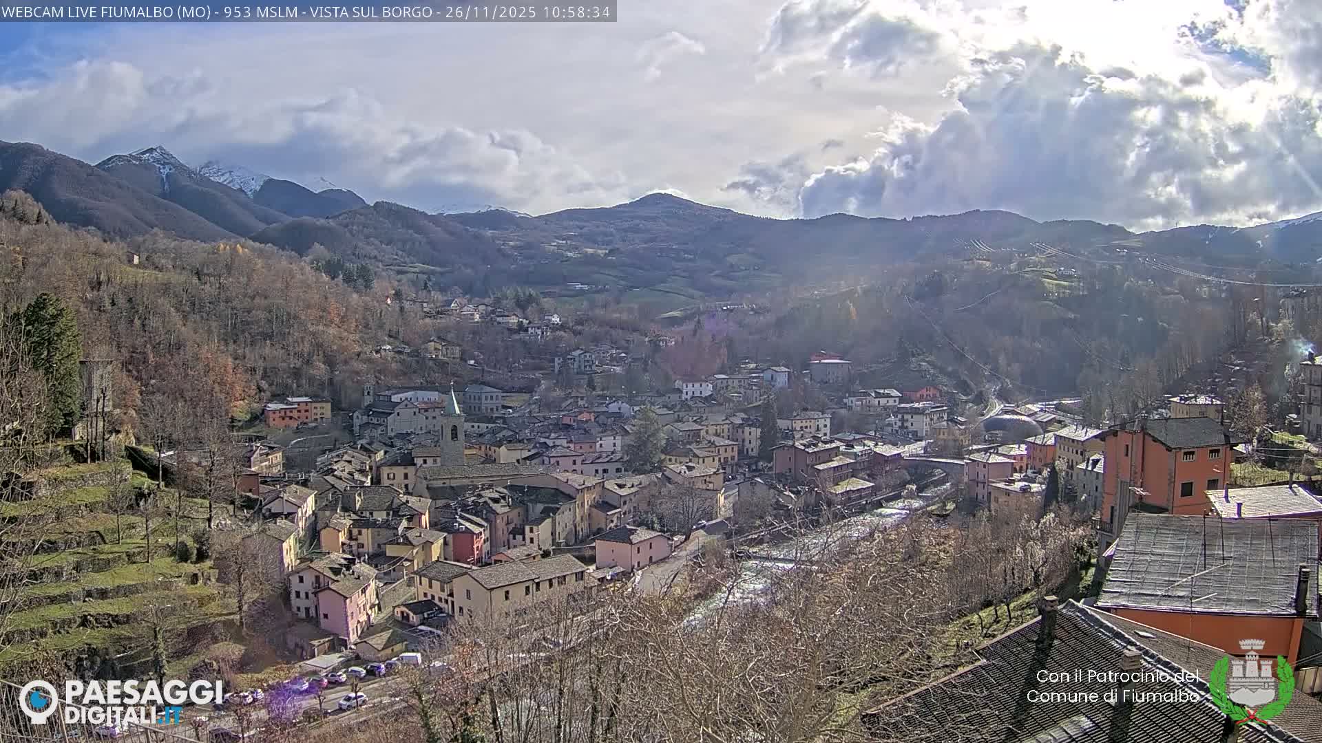 An elevated view showcases a picturesque European town with numerous colorful buildings and a prominent church steeple, nestled in a valley alongside a river, surrounded by hillsides dotted with bare trees and distant snow-capped mountains under a bright, partly cloudy sky.