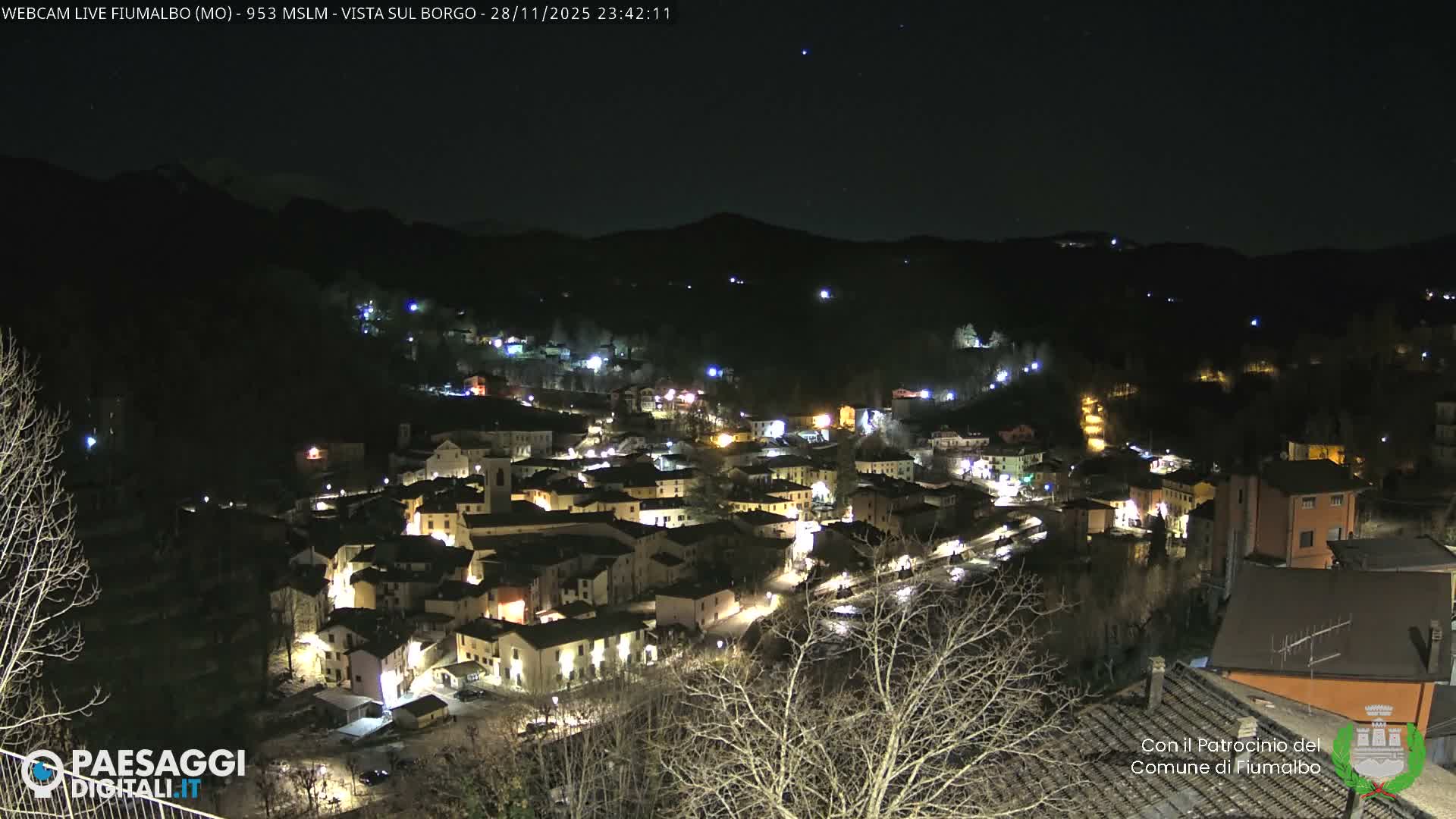 A picturesque mountain village glows warmly with numerous lights at night, nestled among dark hills and bare trees under a clear, starry sky.
