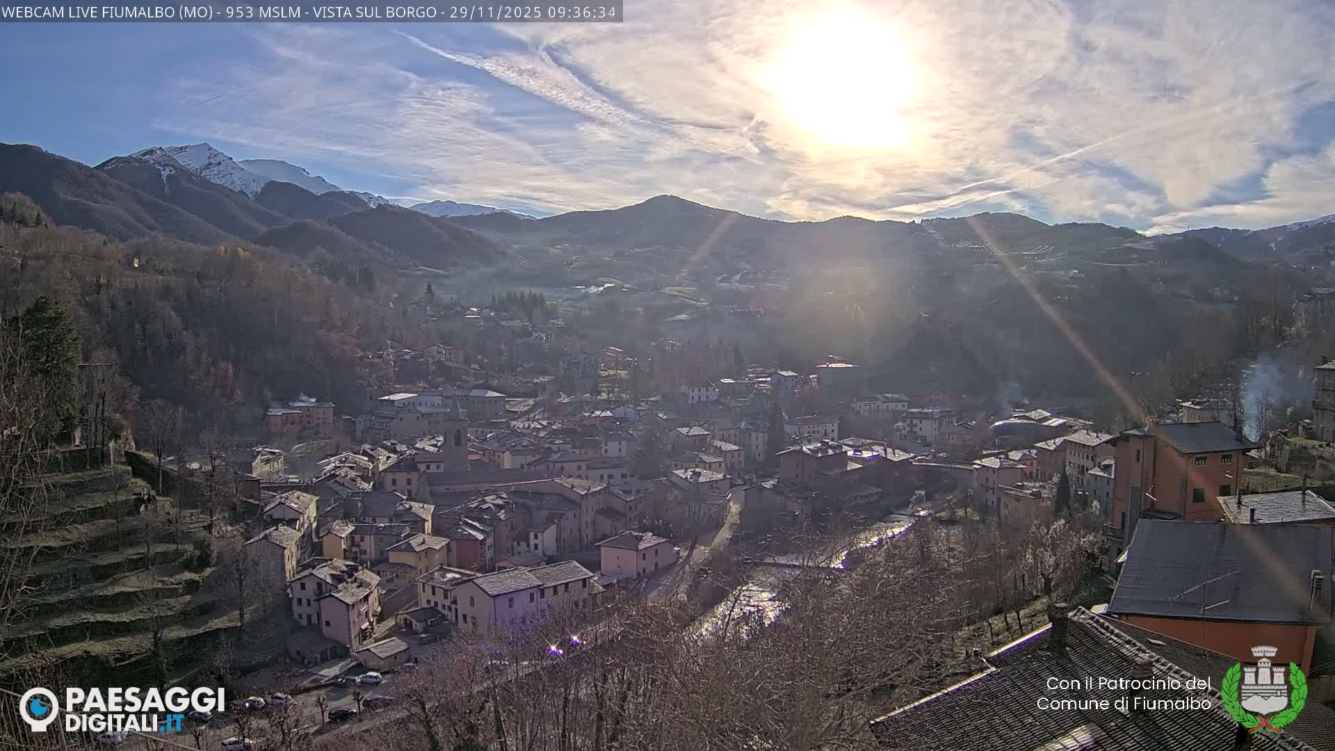 A picturesque mountain village with a river winding through it is nestled in a valley, framed by forested hills and distant snow-capped peaks under a bright, partly cloudy sky.