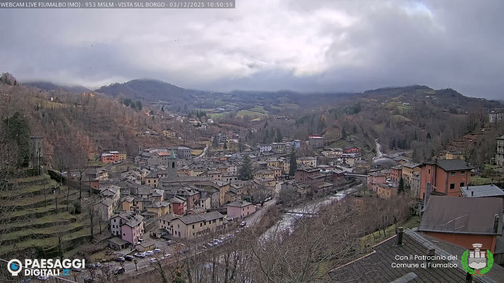 A village with various colored buildings and a river flows through a valley surrounded by mostly bare hills under a grey, cloudy sky.