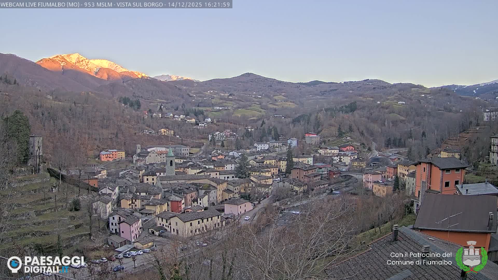 A village with various colored buildings and a river flows through a valley surrounded by mostly bare hills under a grey, cloudy sky.