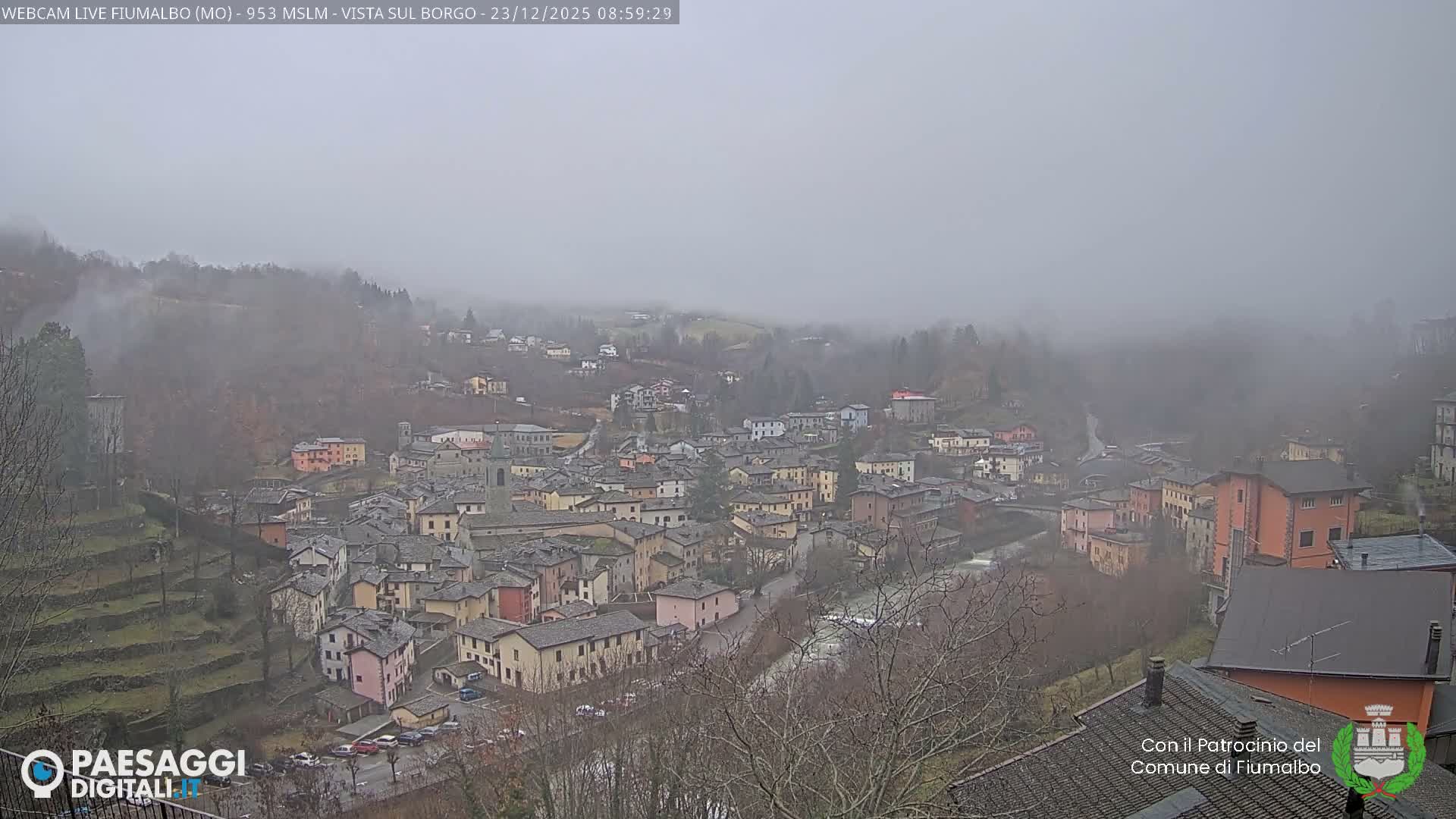 A dense European village with colorful buildings, dark roofs, a church steeple, and a river winding through it is visible amidst terraced hillsides on a very foggy and overcast day.