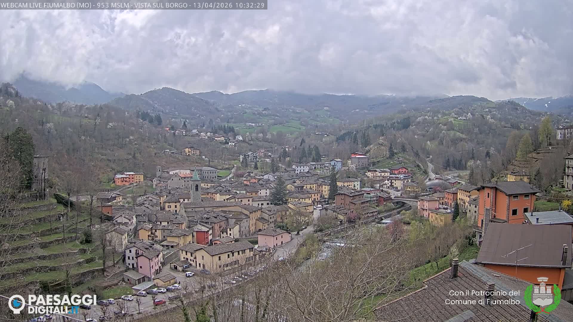 A dense European village with colorful buildings, dark roofs, a church steeple, and a river winding through it is visible amidst terraced hillsides on a very foggy and overcast day.