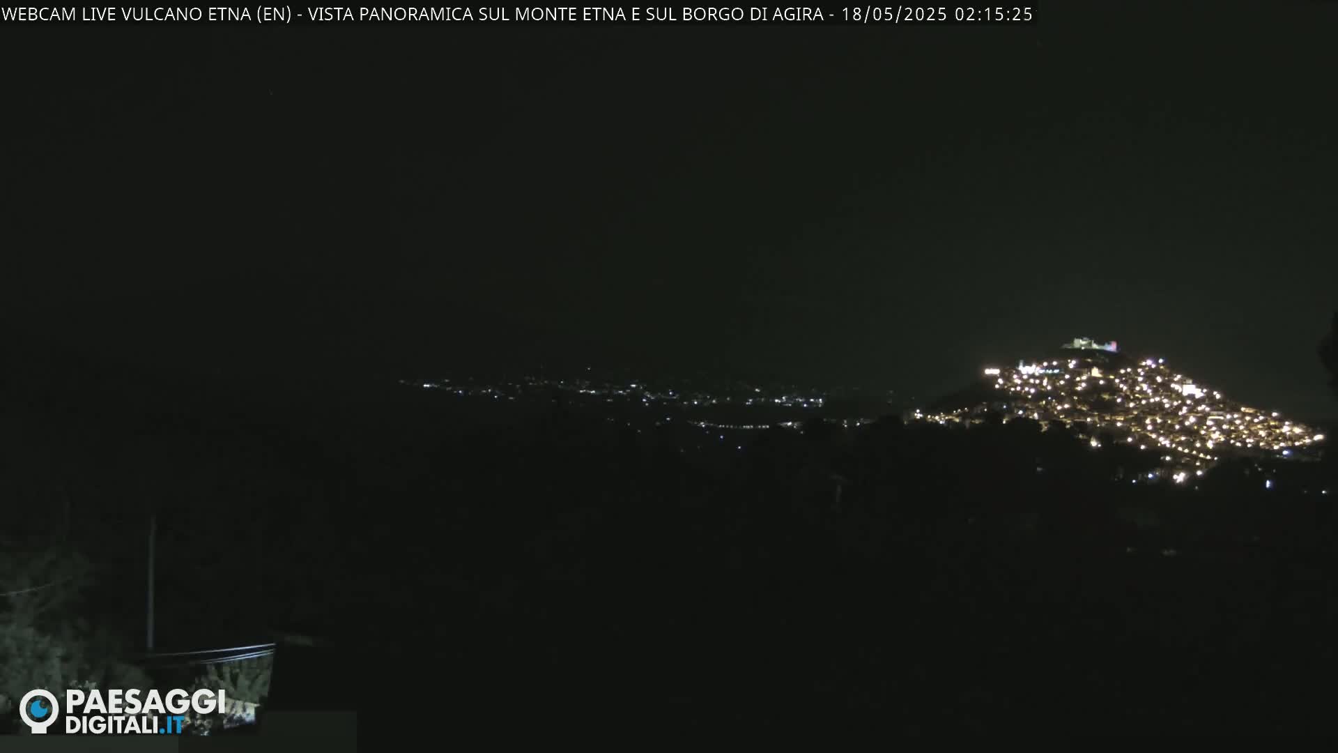 A nighttime, long shot shows a hilltop town illuminated with many lights, set against a dark, clear sky.