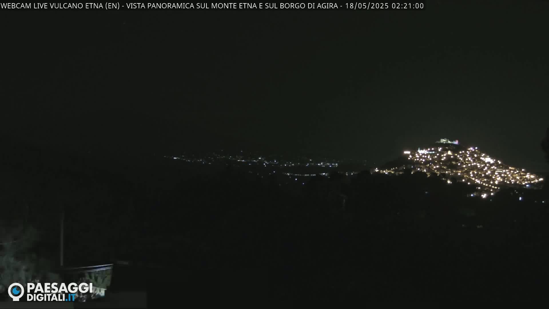 A nighttime long shot shows a hilltop town illuminated with many small lights against a dark, clear sky.