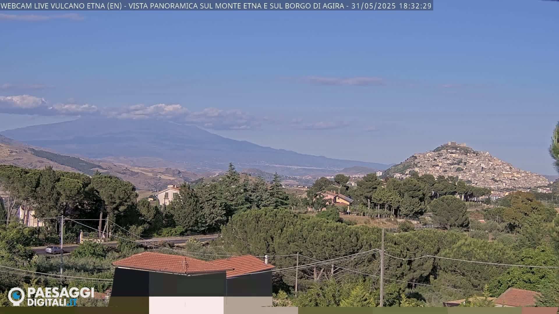 A panoramic view shows a town nestled on a hill, with Mount Etna in the distance under a mostly clear, sunny sky.
