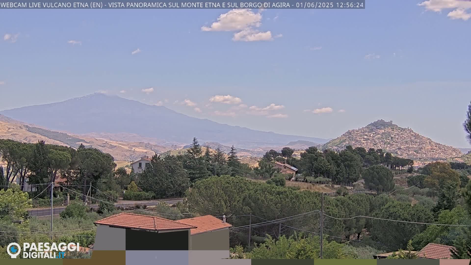 A panoramic view shows a distant, hazy mountain range under a mostly clear sky, with a hilltop town nestled in the foreground amongst green trees and houses.