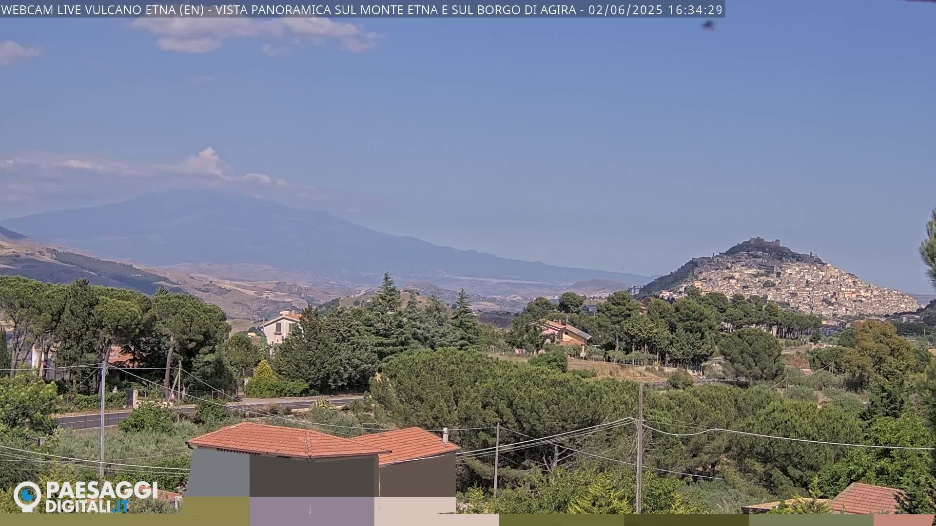 A sunlit view of a town nestled on a hill, with Mount Etna in the background under a mostly clear sky.