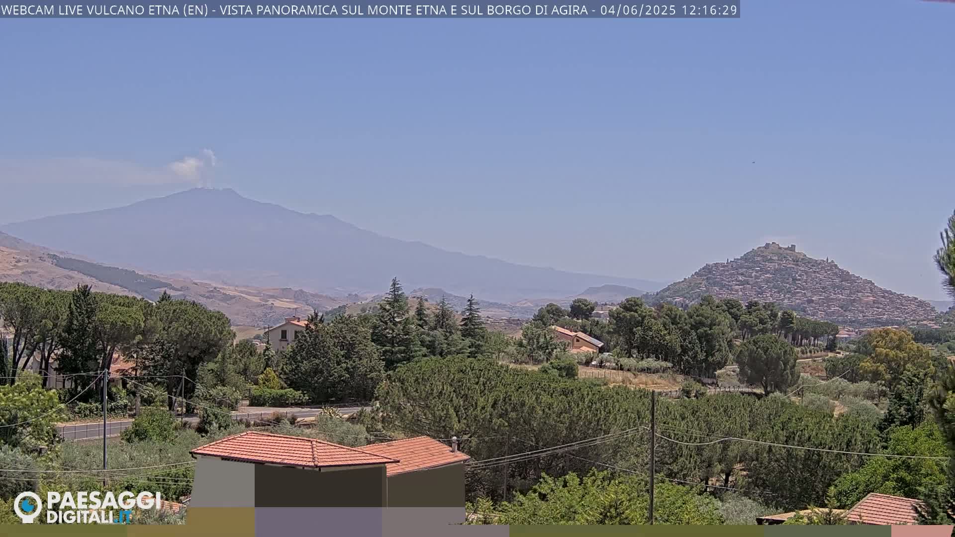 A hazy, sunny day reveals a distant, smoking Mount Etna, a hilltop town, and a foreground of green trees and houses.