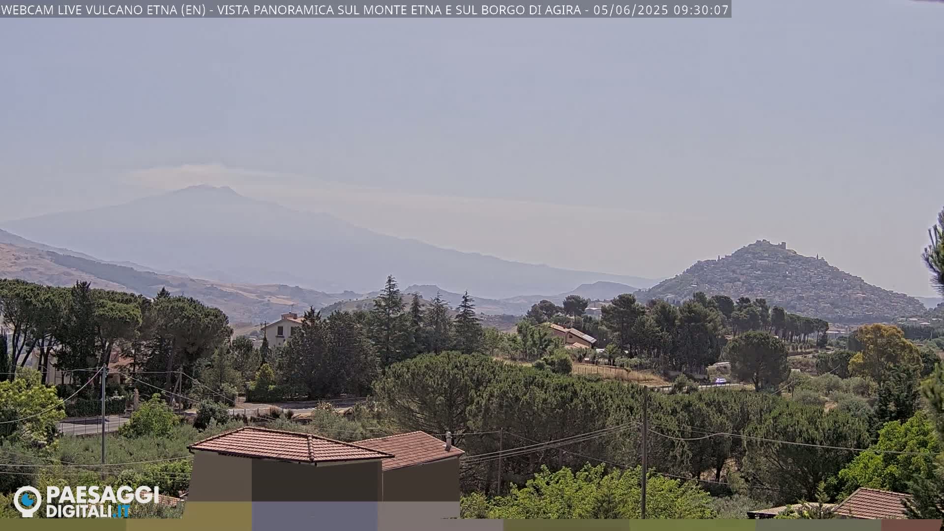 A hazy view shows a hilltop town nestled among green trees and rolling hills, with a distant, hazy mountain range under a clear sky.
