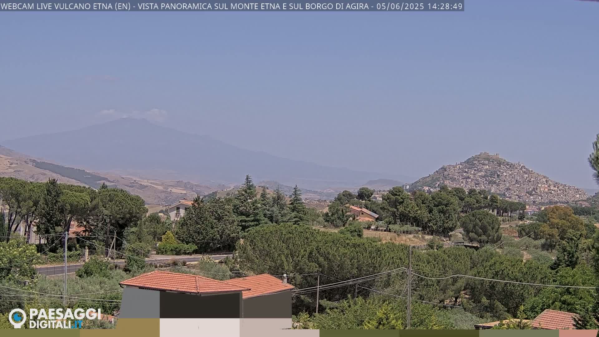 A hazy, sunny day reveals a panoramic view of Mount Etna in the distance, a hilltop town nestled in the foreground, and a verdant landscape filled with trees and buildings.