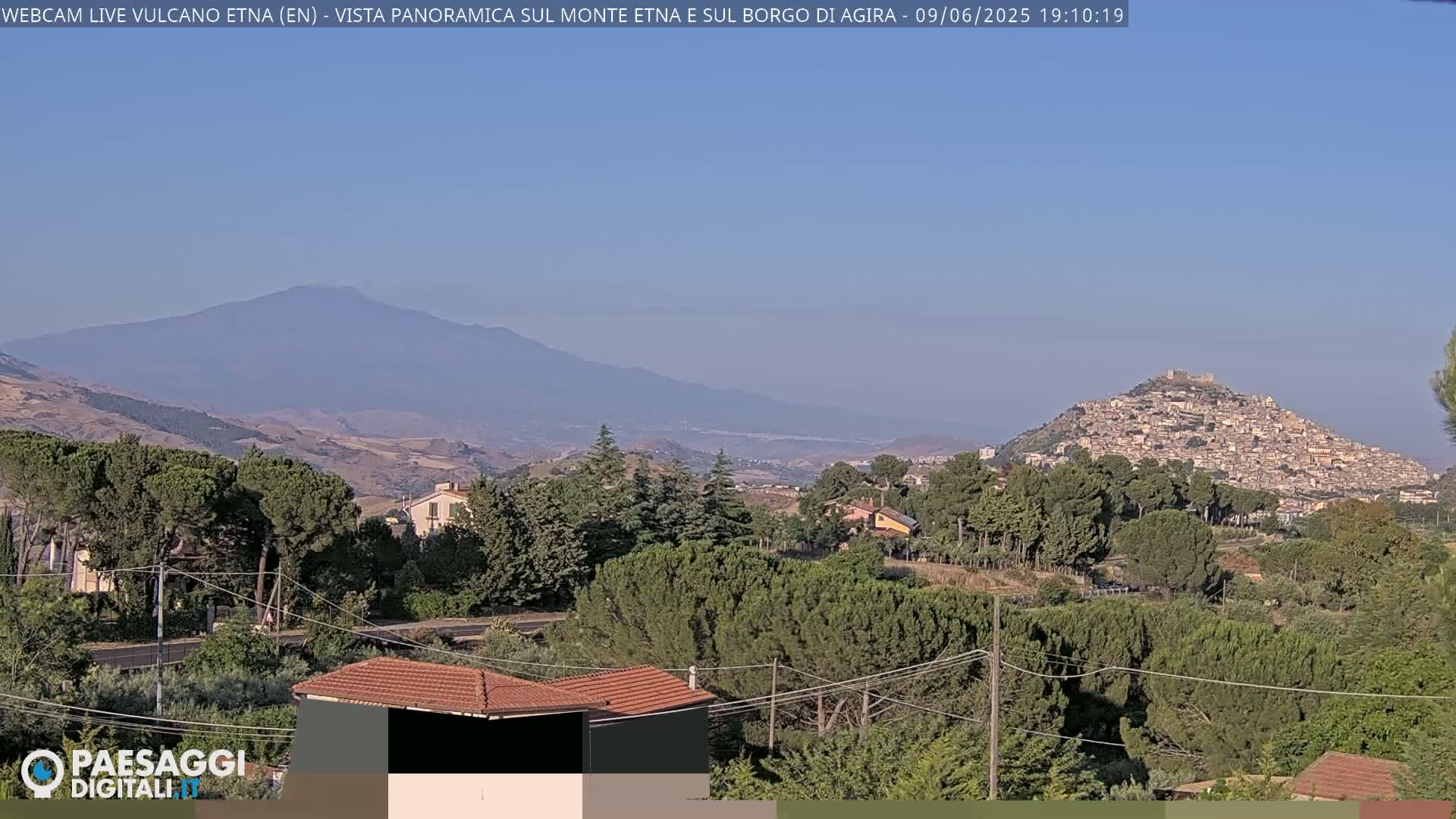 A distant, hazy view of Mount Etna and a hilltop town, under a clear, light-blue sky, is seen from a vantage point amid green, leafy trees and a few houses.