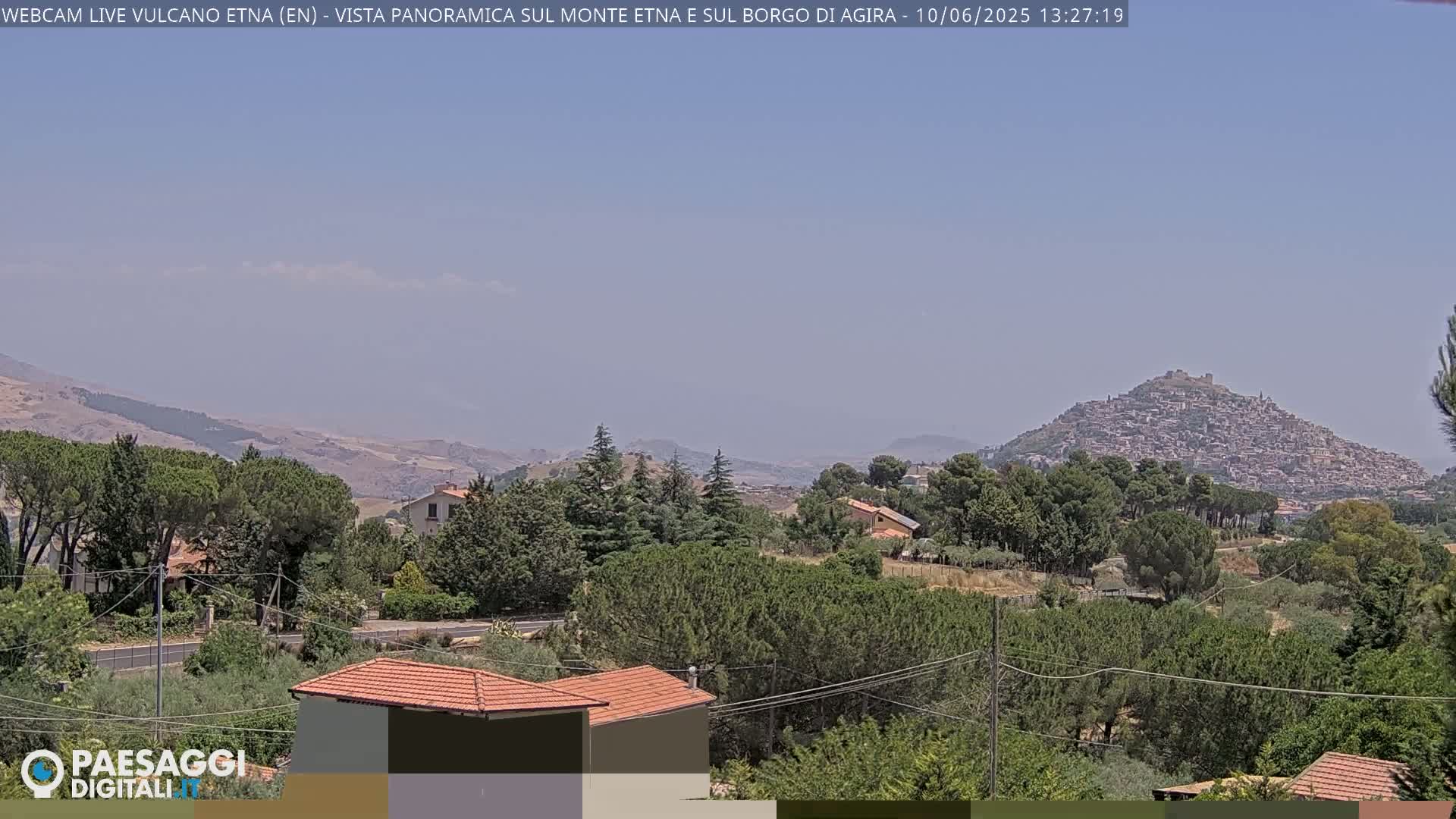 A hazy, sunny day reveals a panoramic view of a hilltop town nestled among green hills and trees, with mountains in the far distance.