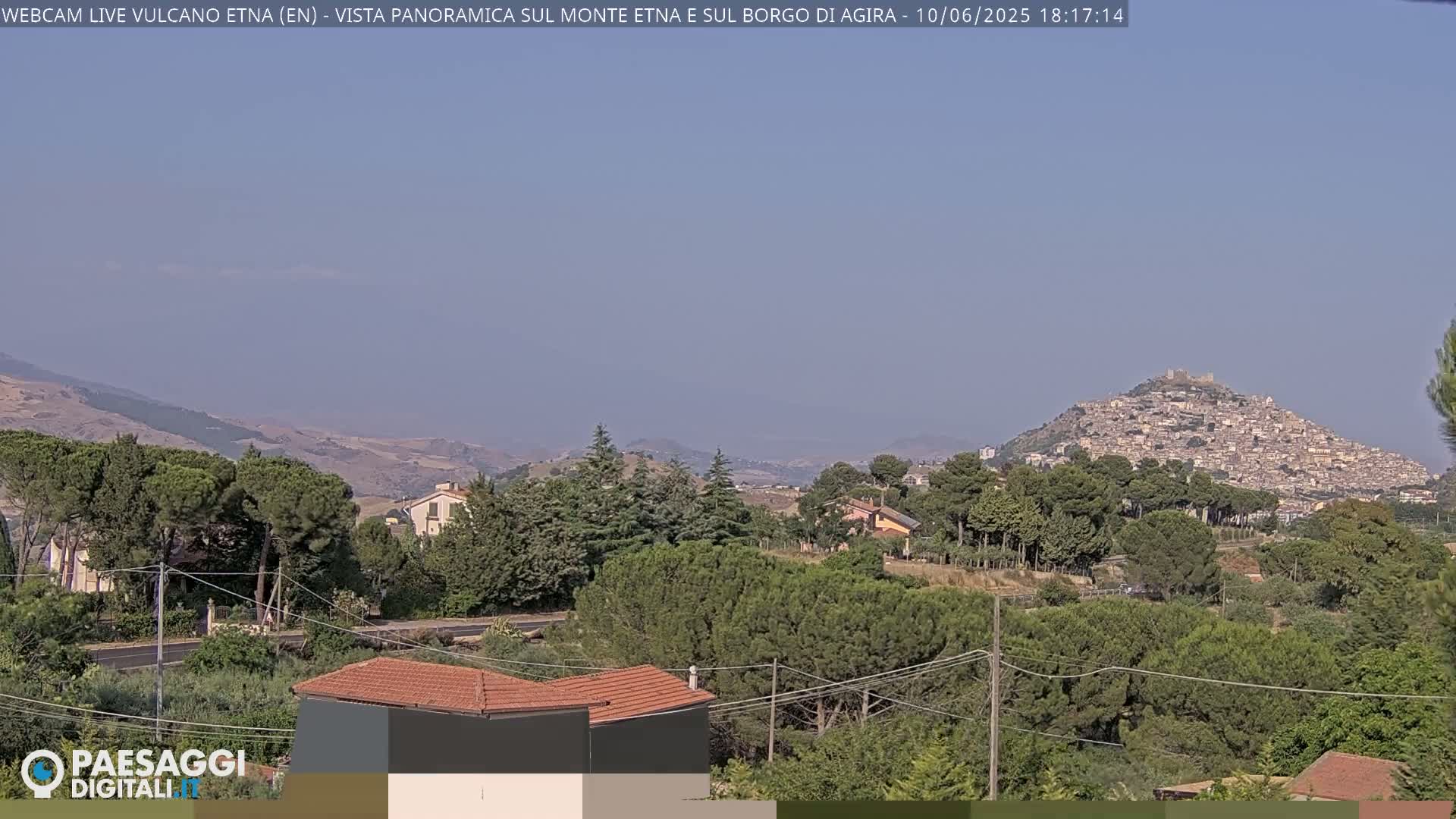 A hazy, sunny day reveals a hilltop town nestled among green hills and trees, with distant mountains visible in the background.