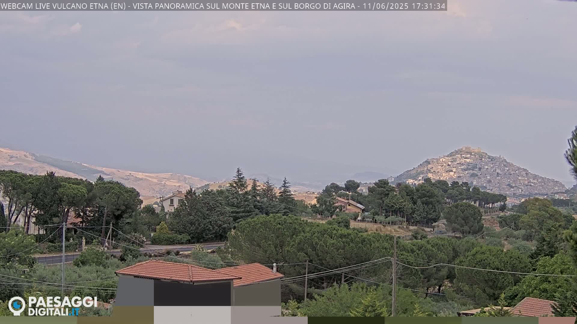A hilltop town is visible in the distance, under a hazy sky, with lush green vegetation and several houses in the foreground.