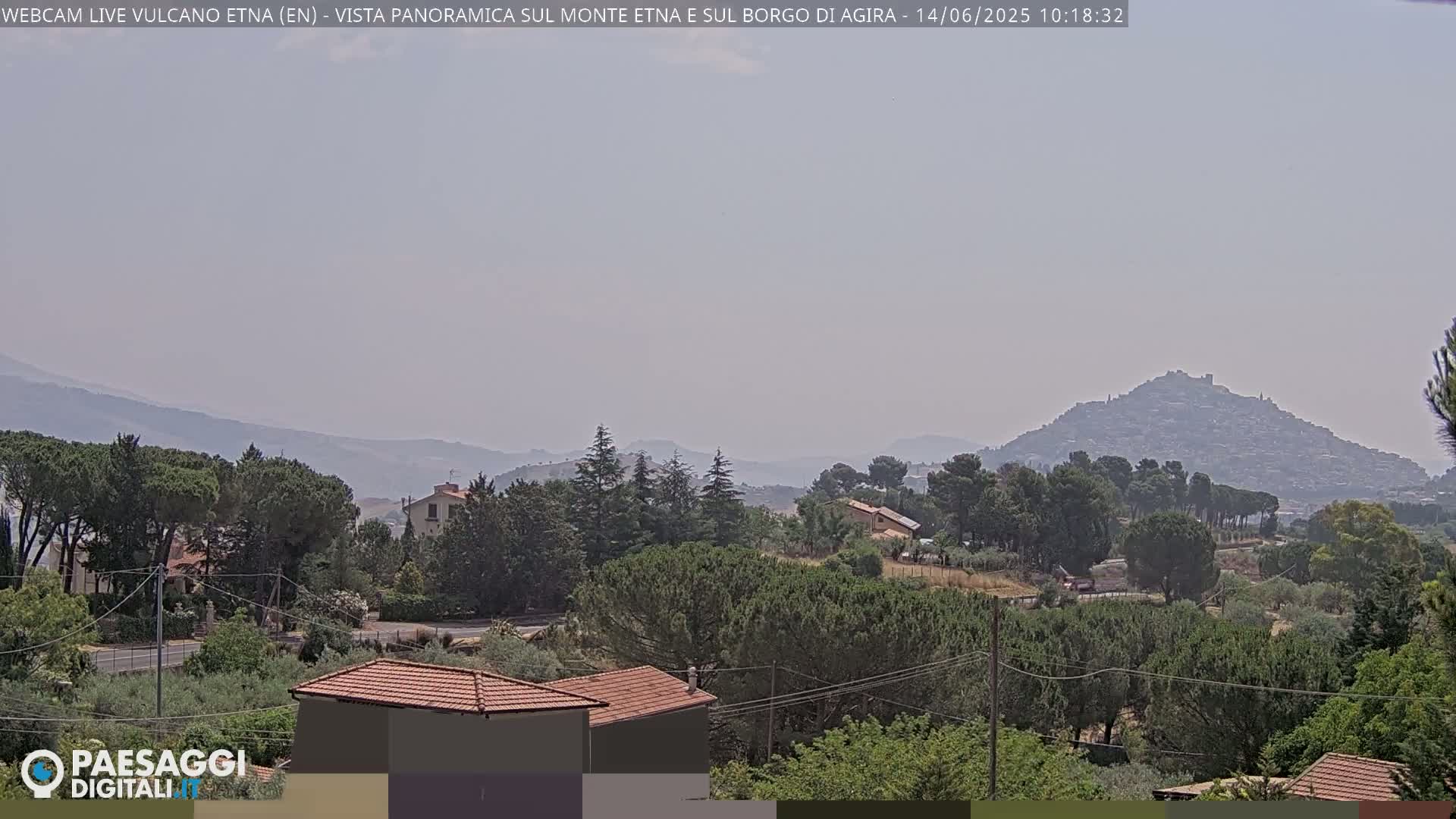 A hazy, sunny view of a hilltop town nestled among green, tree-covered hills and low-lying houses.