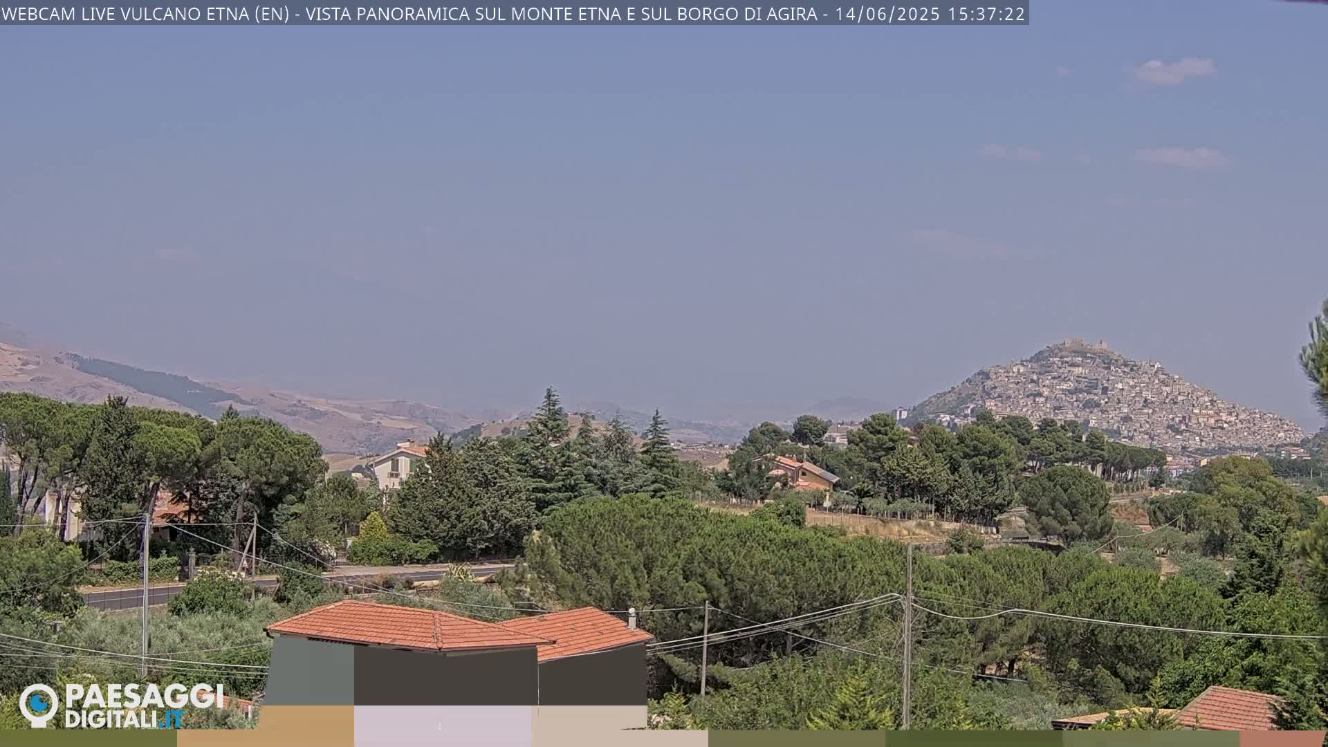 A hazy, sunny day reveals a panoramic view of a town nestled atop a hill, surrounded by lush green vegetation and rolling hills in the distance.
