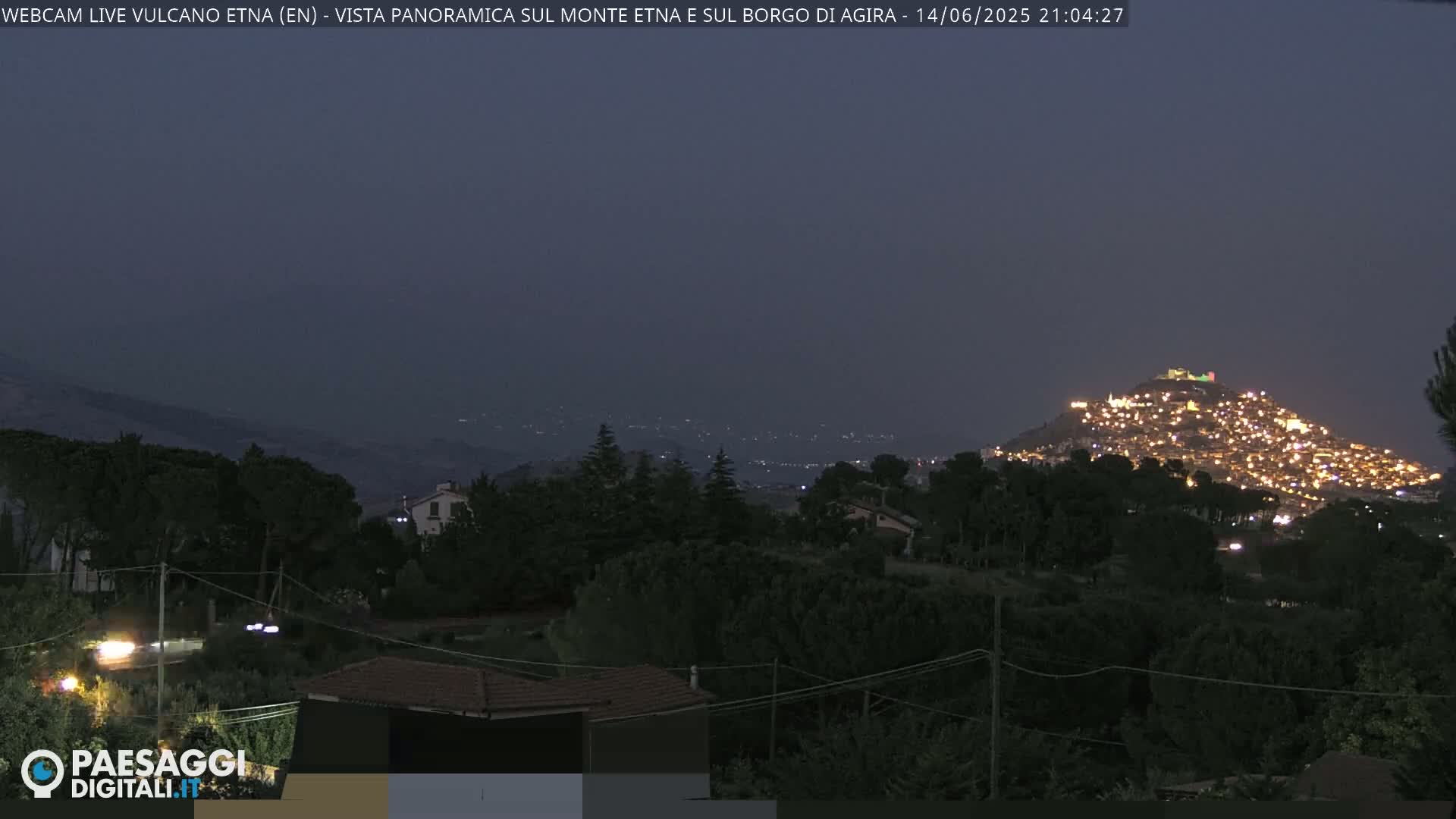 A hazy, sunny day reveals a panoramic view of a town nestled atop a hill, surrounded by lush green vegetation and rolling hills in the distance.