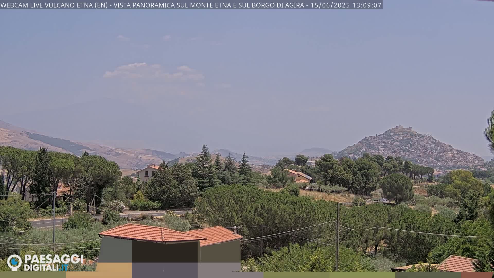 A panoramic view on a partly cloudy day shows a town nestled on a hill, surrounded by lush green vegetation and distant mountains.