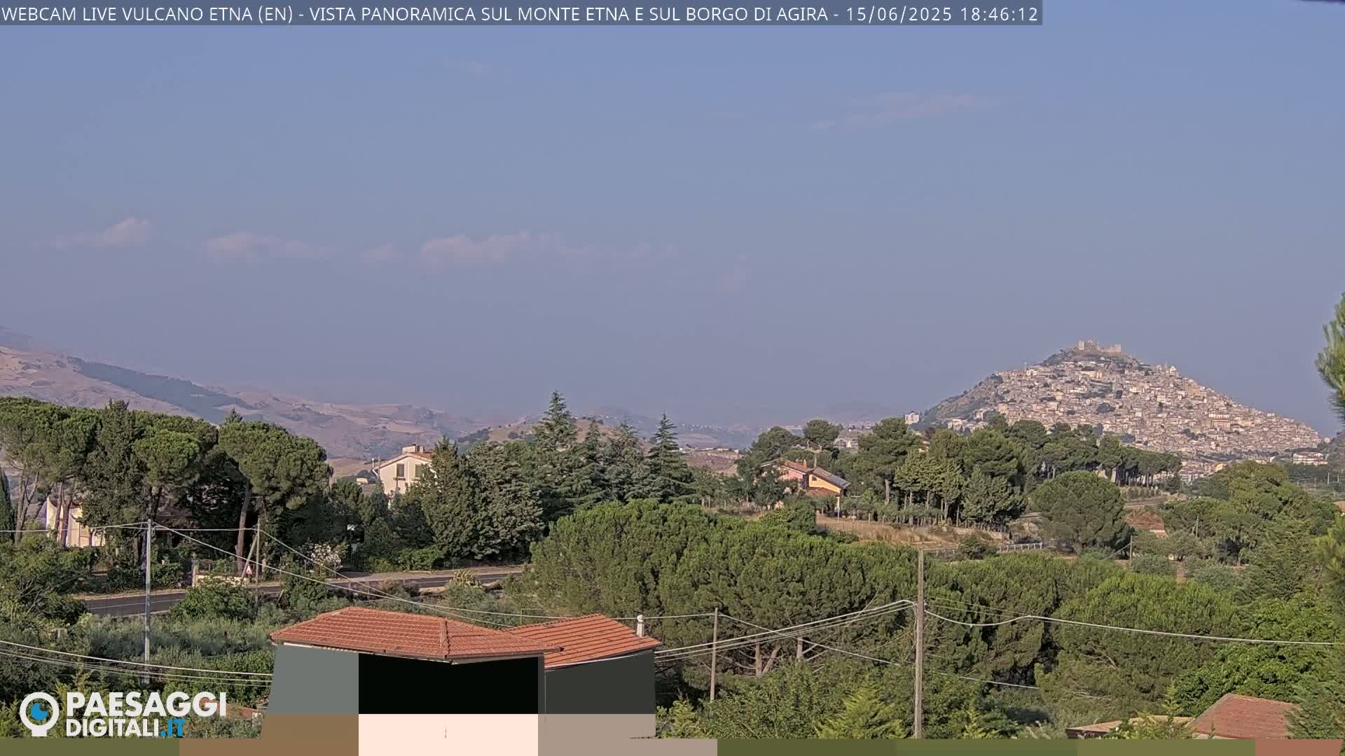 A hazy, sunny day reveals a hilltop town nestled among green trees and rolling hills, with a distant mountain range under a pale blue sky.