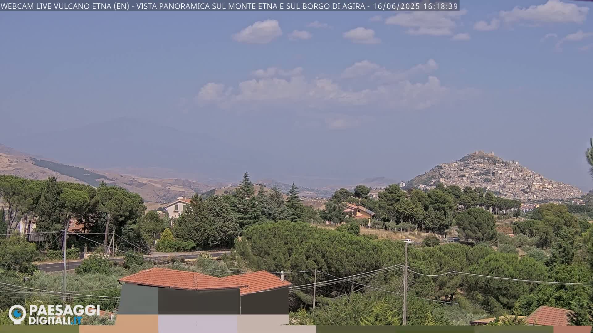 A partly cloudy day reveals a panoramic view of a hilltop town nestled amongst green trees and rolling hills, with a mountain range in the far distance.