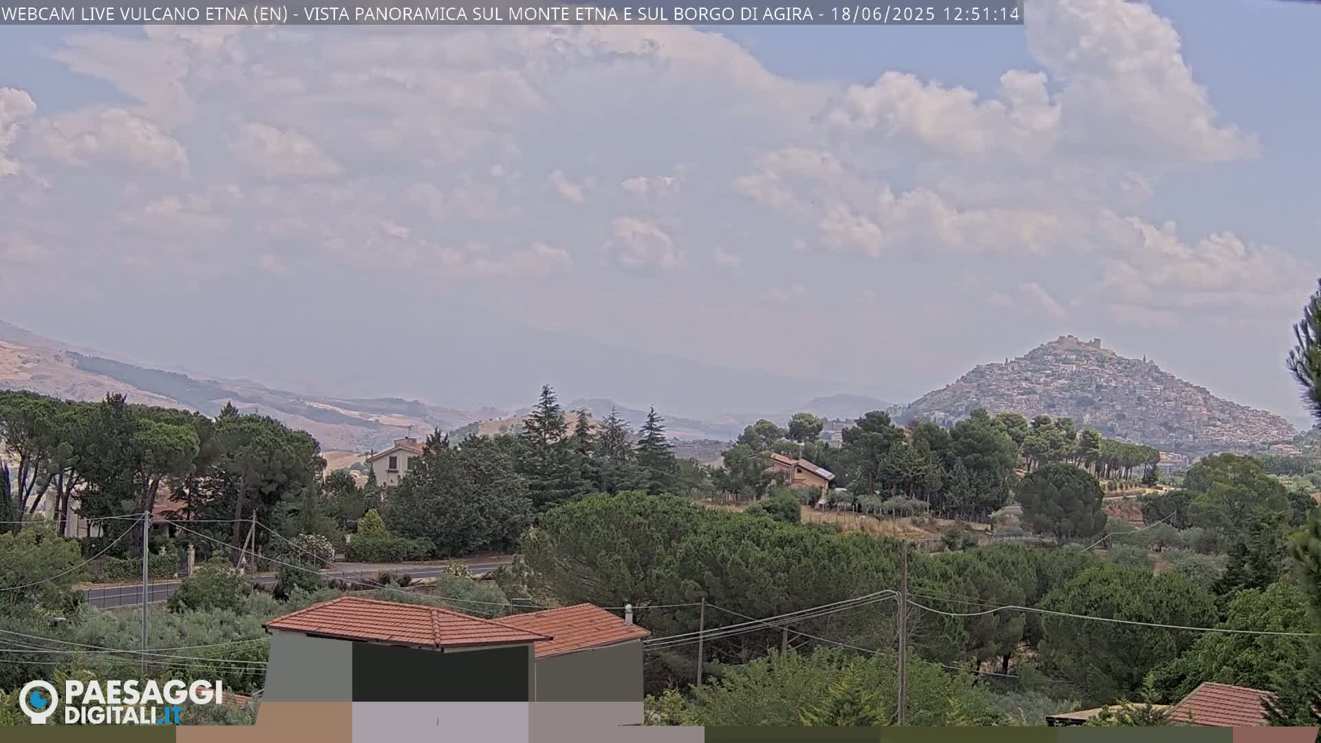 A partly cloudy day reveals a view of a hilltop town nestled among green trees and rolling hills, with mountains in the distance.