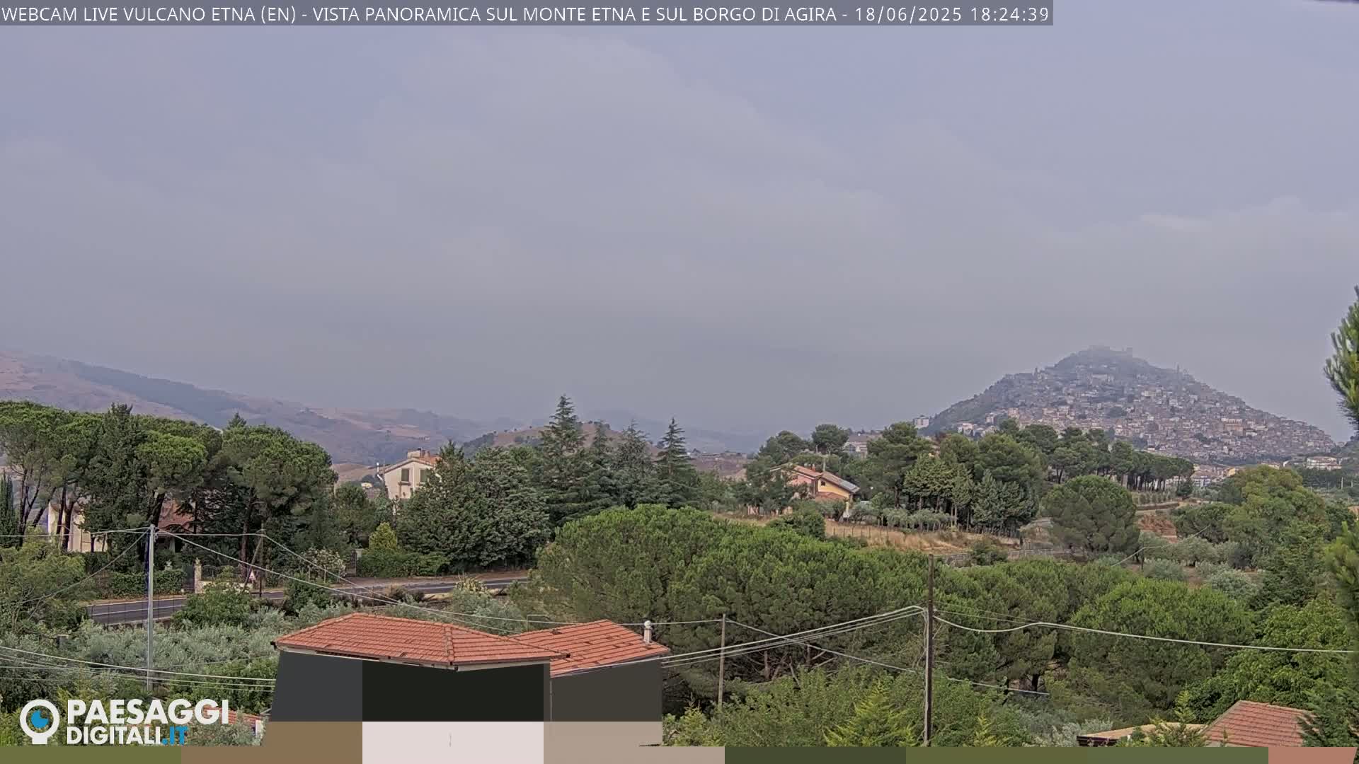 A hazy, long shot shows a town nestled on a hill, surrounded by green trees and shrubs, with more hills and mountains visible in the distance.