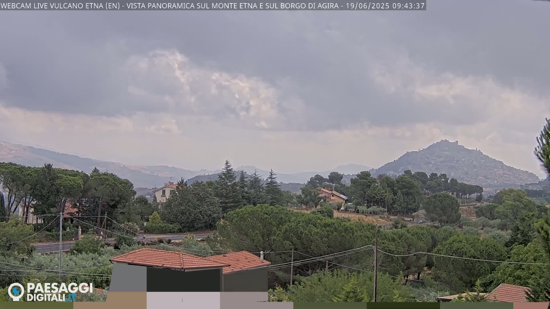 A panoramic view of a hilly landscape under an overcast sky, showing a town nestled on a distant hill among green trees and houses.