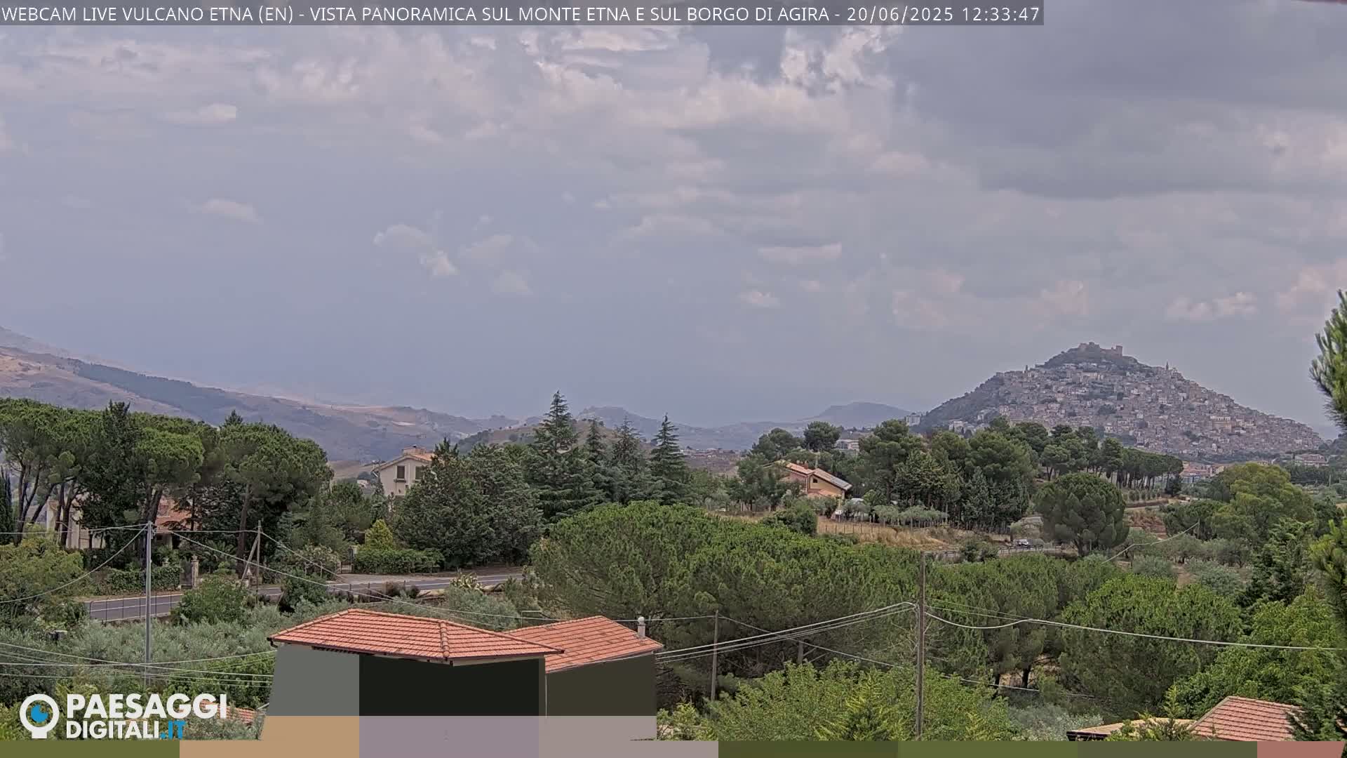 A panoramic view of a town nestled on a hill, surrounded by green trees and rolling hills under a partly cloudy sky.