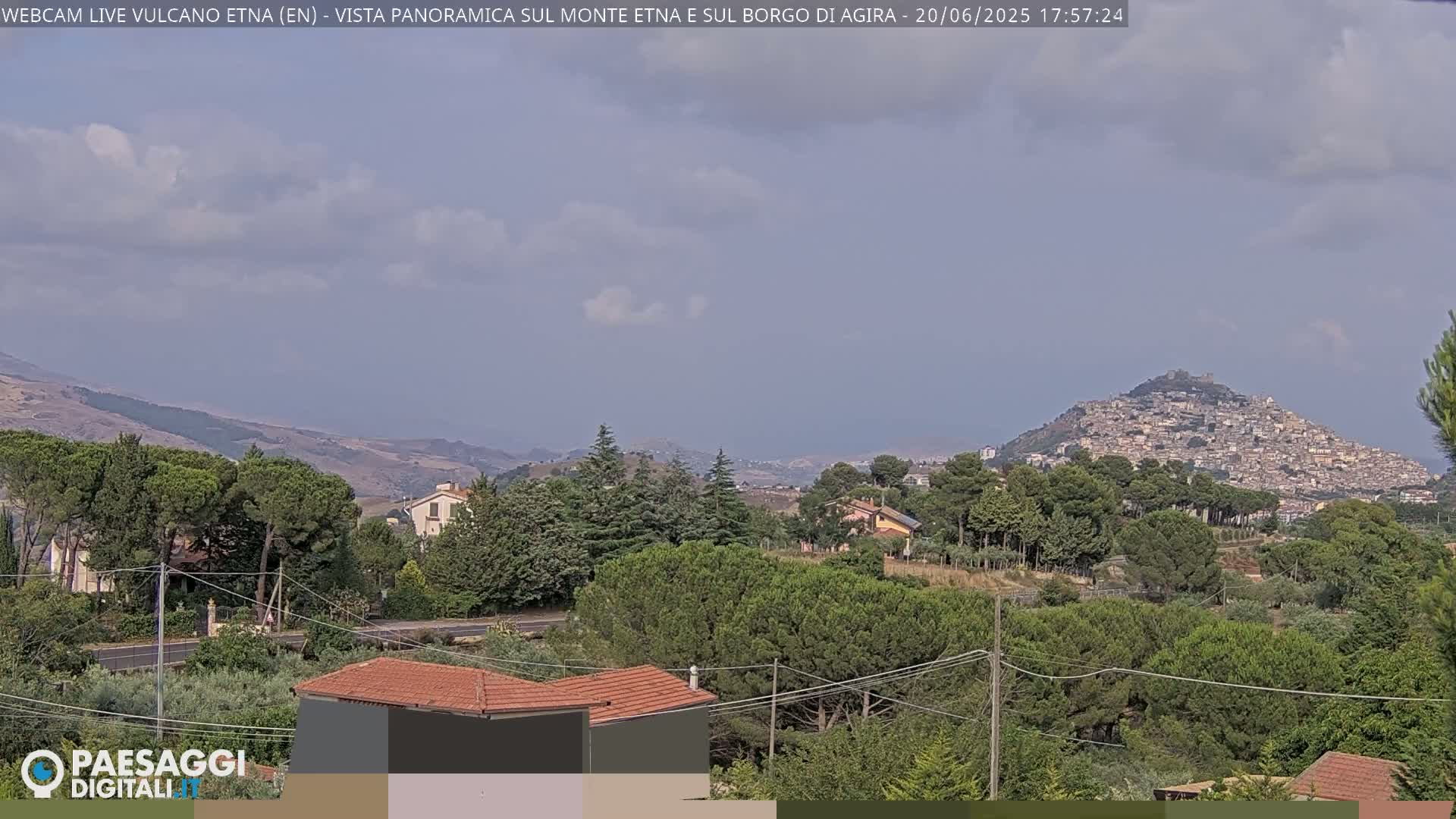 A partly cloudy day reveals a hilltop town nestled among green trees and rolling hills, with other houses and buildings visible in the foreground.