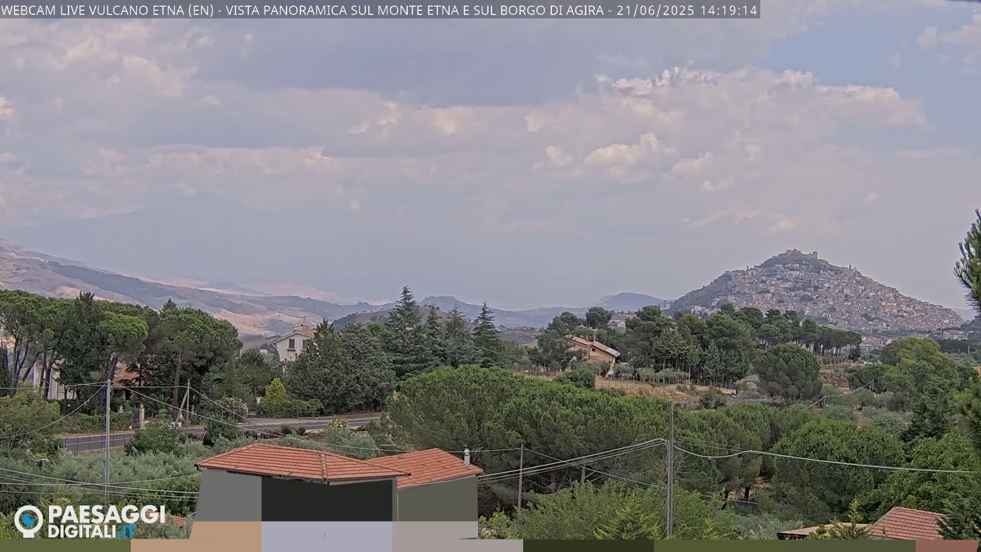 A partly cloudy day reveals a panoramic view of a hilltop town nestled among green trees and rolling hills.