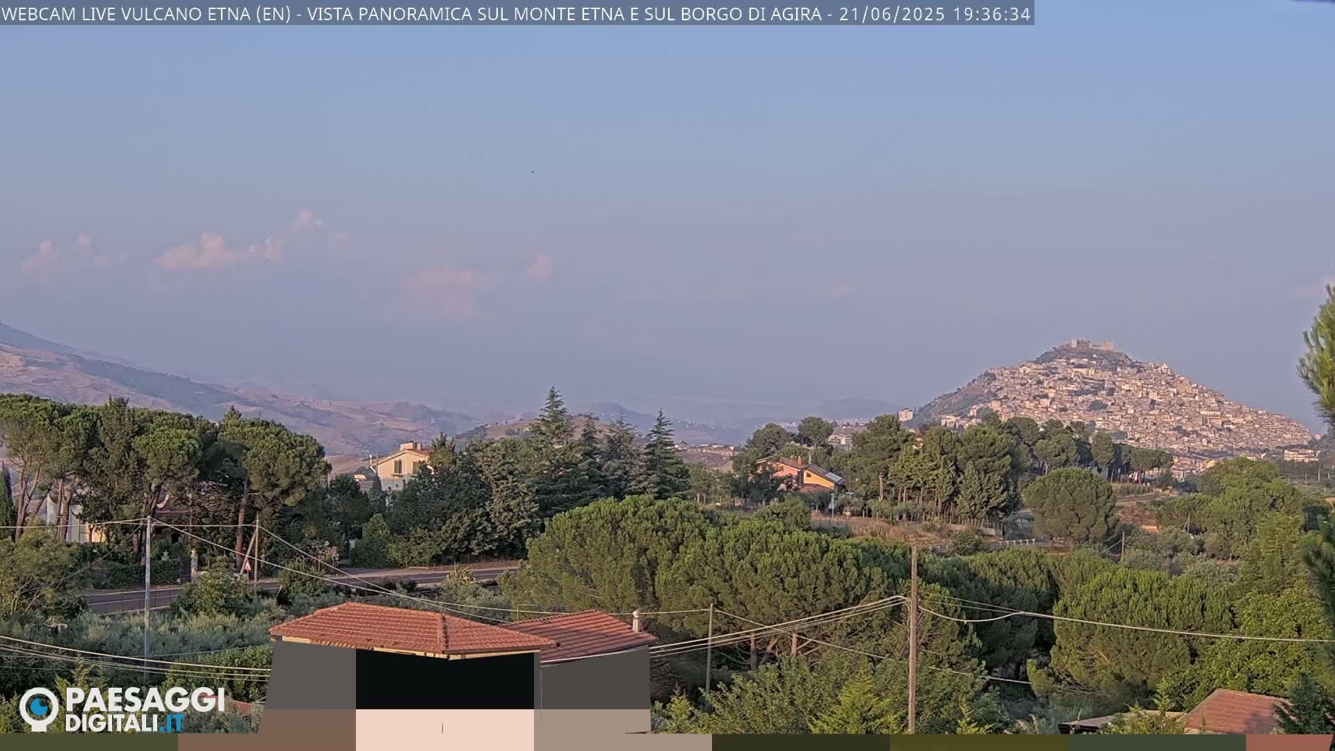 A hazy, sunny day reveals a hilltop town nestled among green trees and rolling hills, with mountains in the background.