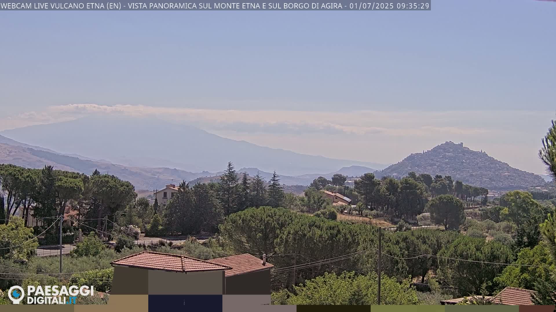 A hazy, sunny day reveals a panoramic view of a mountain range, a hilltop town, and a verdant landscape with houses nestled among the trees.