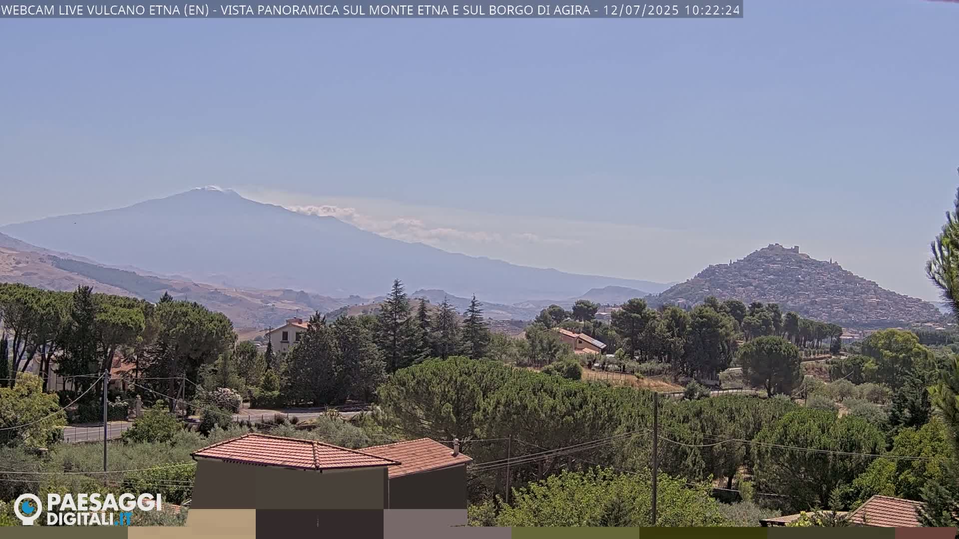 A distant, snow-capped mountain is visible under a clear, light blue sky, behind a hilltop town and a foreground of green trees and houses.