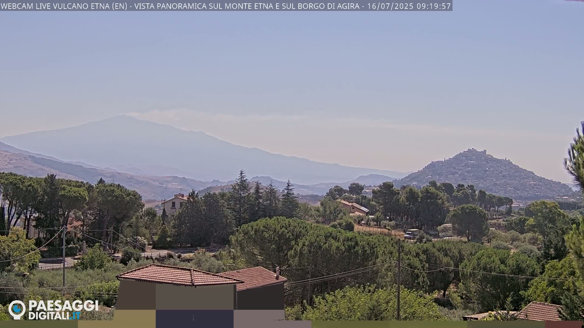 A hazy, mountainous landscape features a distant volcano, a hilltop town, and a foreground of green trees and houses under a clear sky.