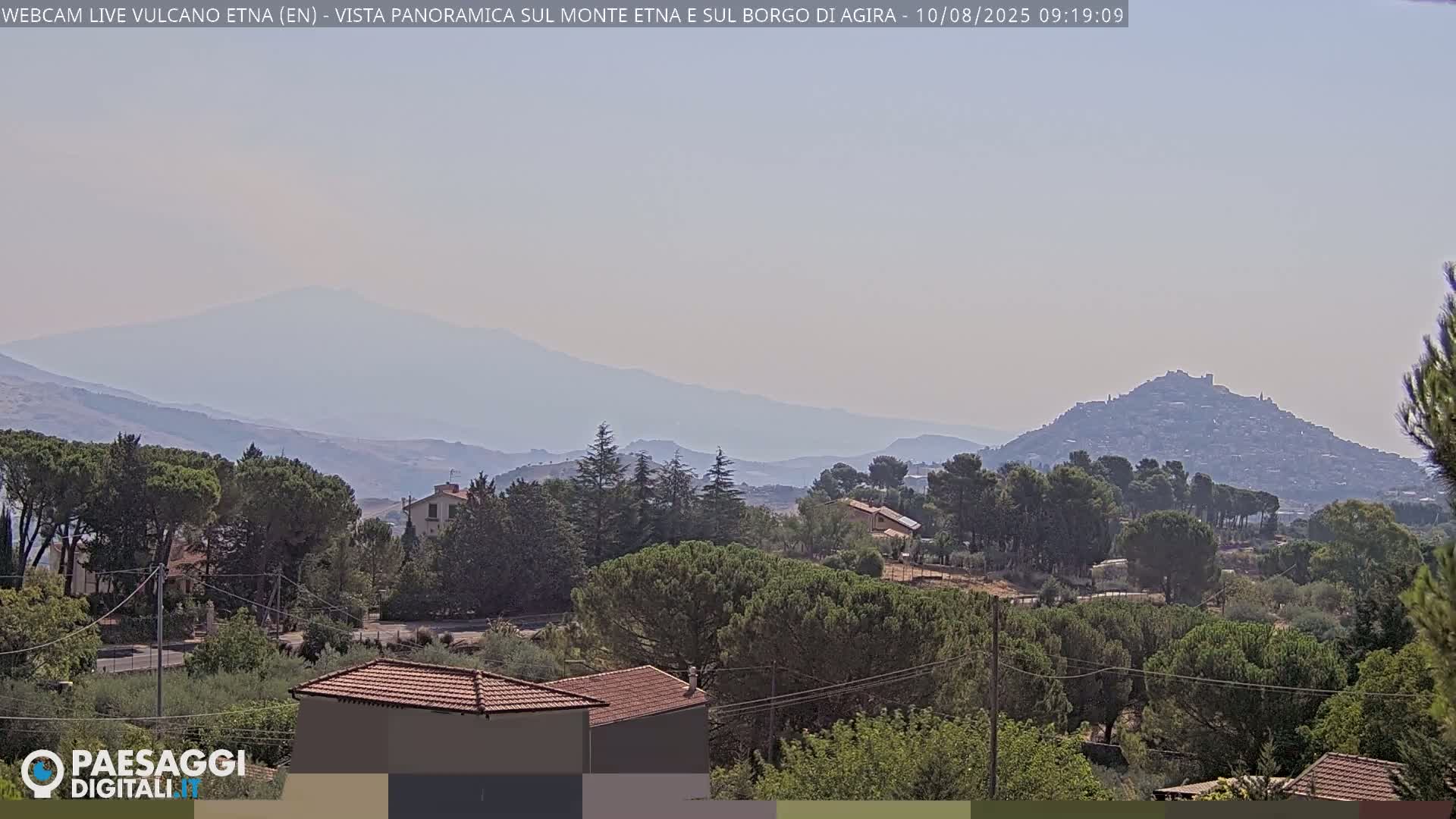 A hazy, long-distance view shows a town nestled on a hill, with additional hills and a mountain range in the background under a mostly clear sky.