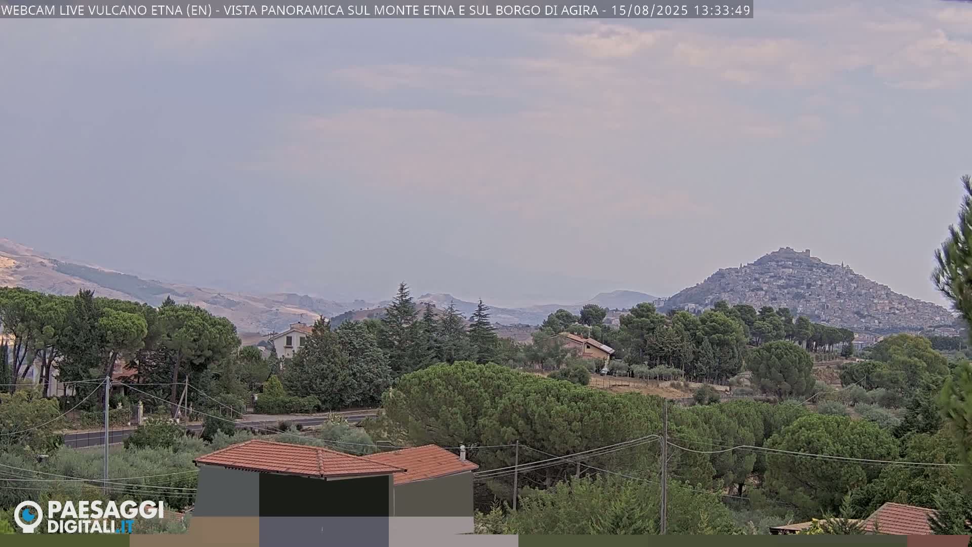 A hazy, long-range view shows a distant, large mountain range behind a hilltop town and a foreground of houses and trees.