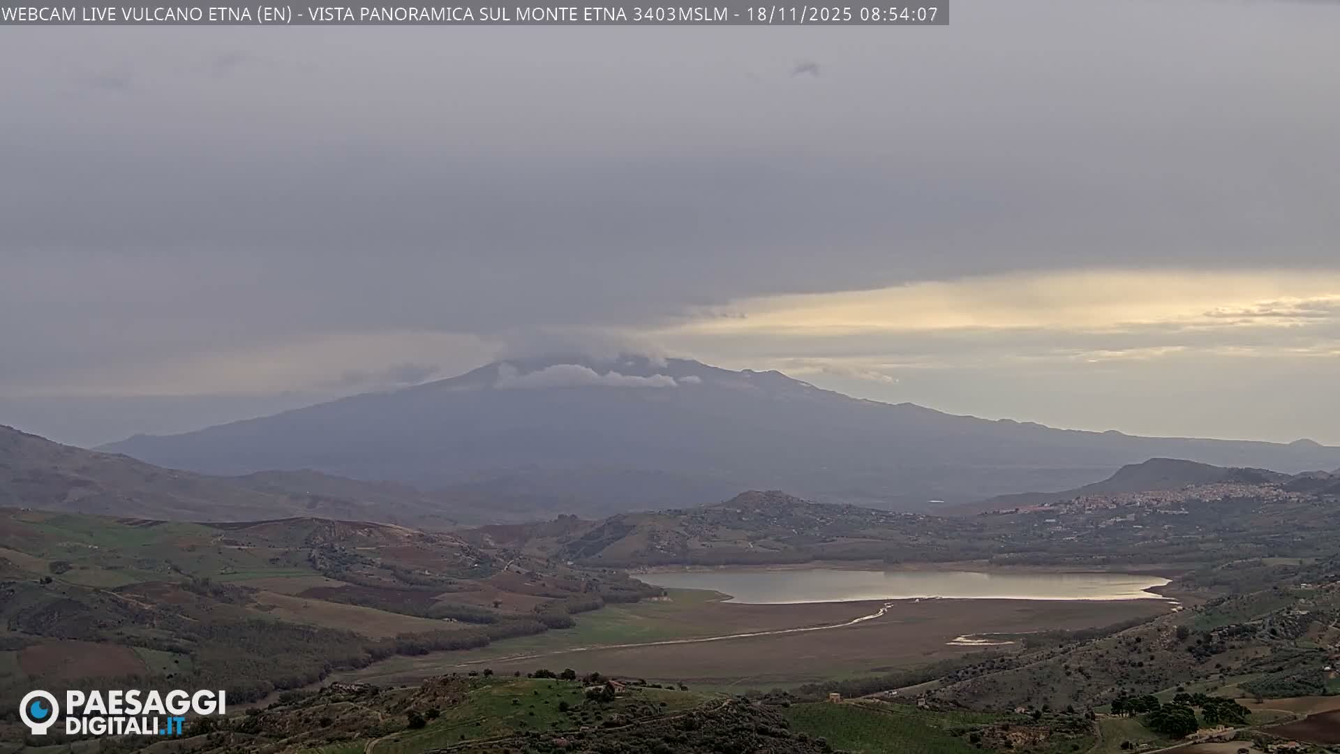 A hazy, long-range view shows a distant, large mountain range behind a hilltop town and a foreground of houses and trees.