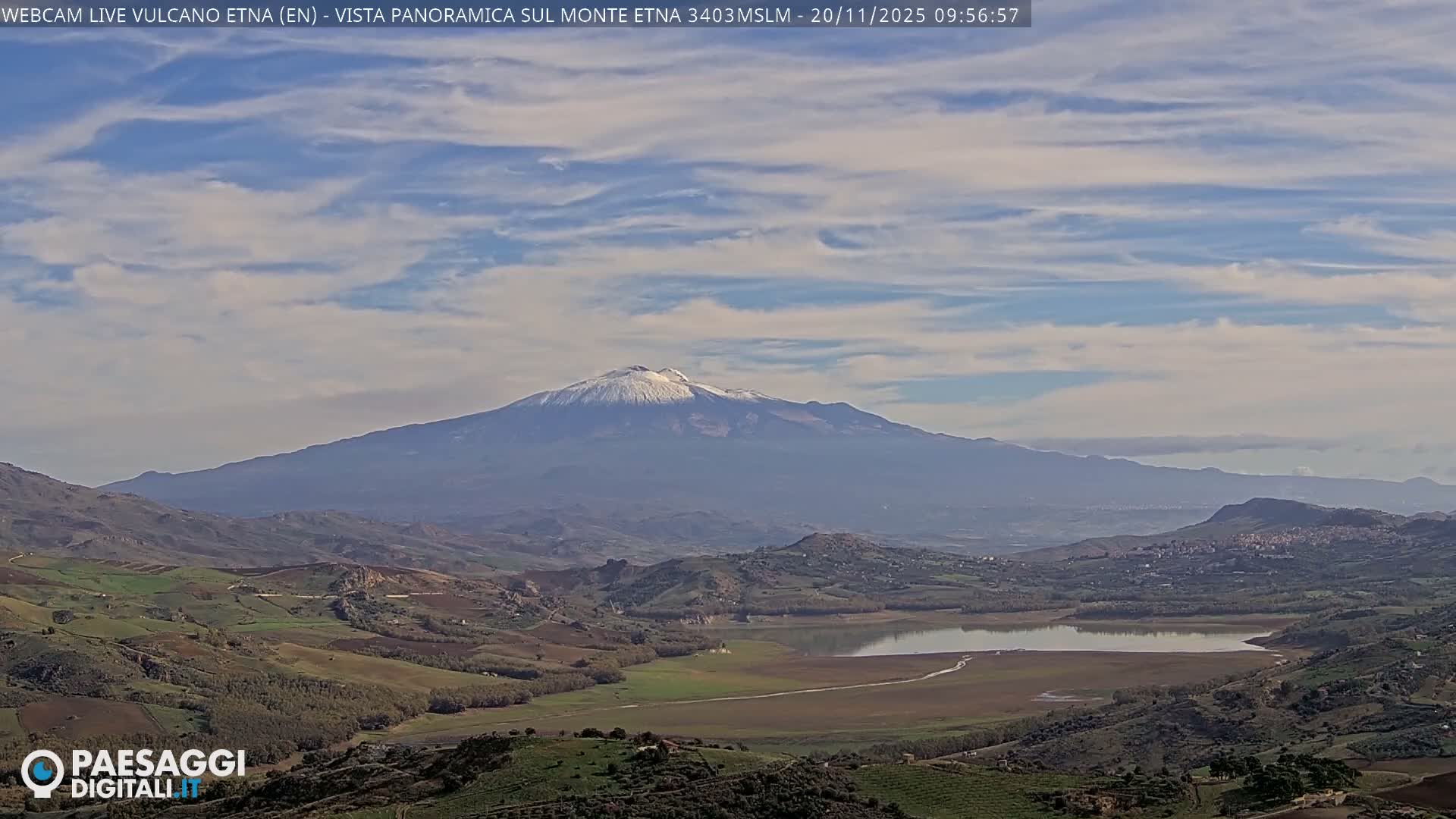 A panoramic view reveals a majestic snow-capped volcano under a partly cloudy sky, overlooking rolling green and brown hills, a winding body of water, and scattered towns.