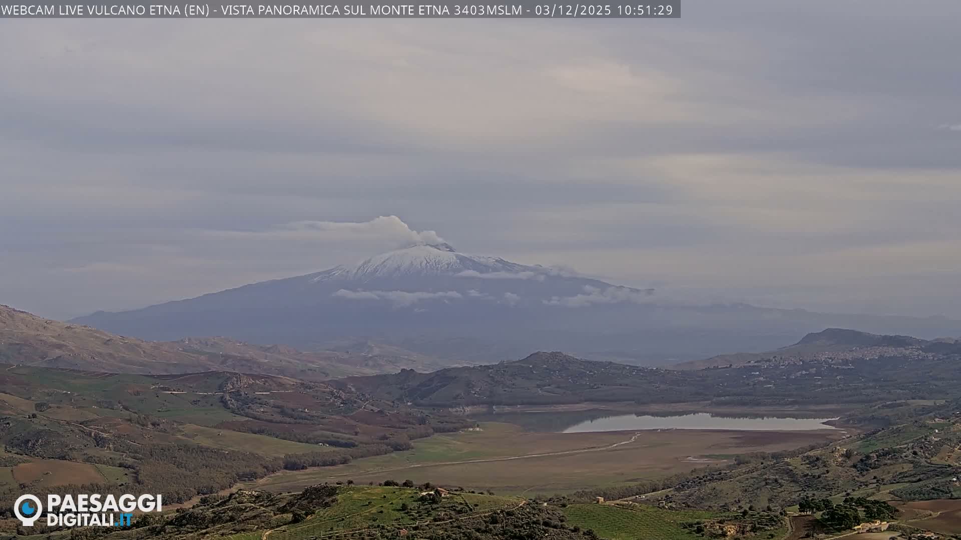 A panoramic outdoor scene under an overcast sky features a snow-capped mountain partially shrouded in clouds, overlooking a valley containing a calm lake, surrounded by rolling green and brown hills with hints of a distant town.