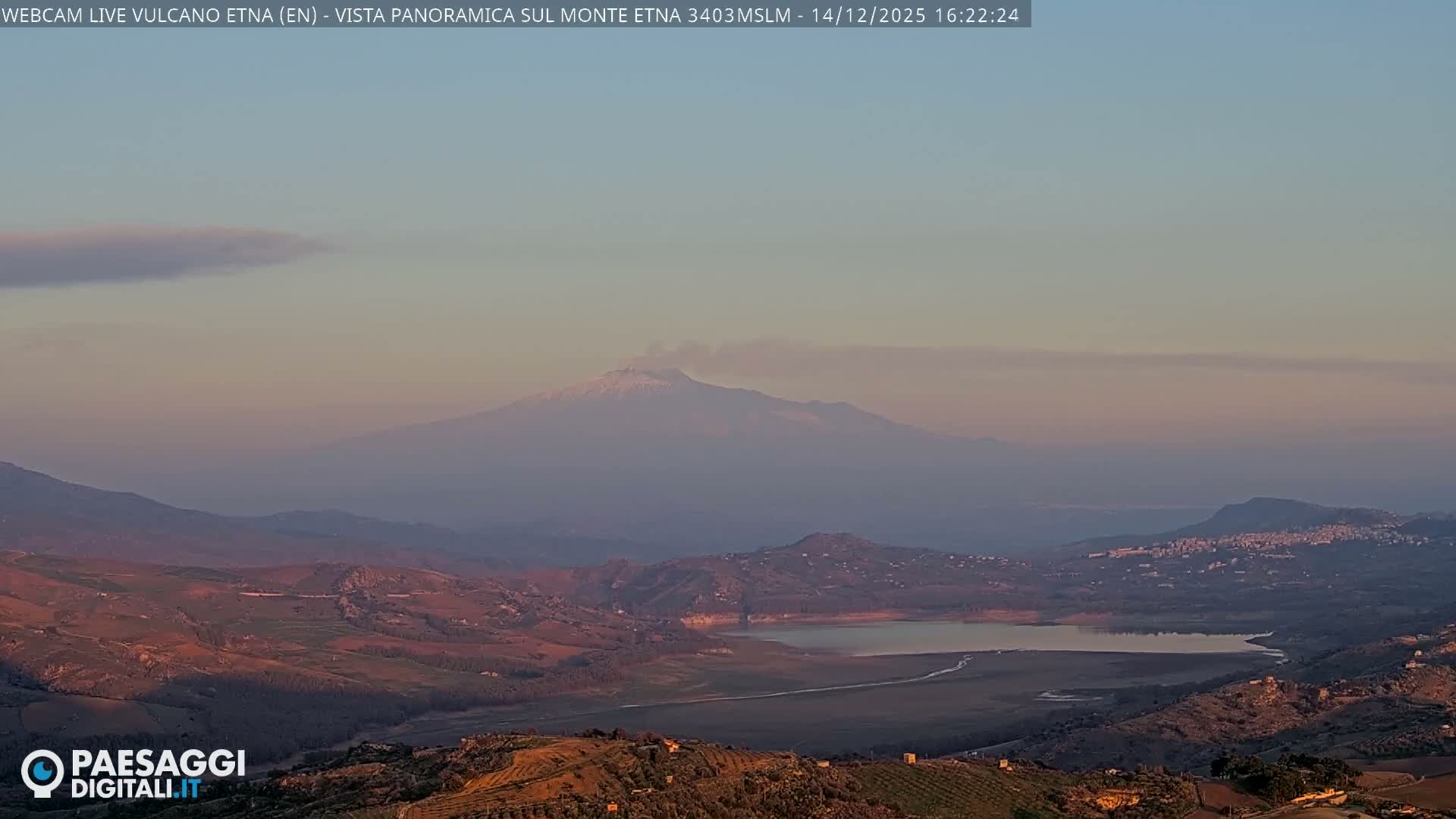 A panoramic outdoor scene under an overcast sky features a snow-capped mountain partially shrouded in clouds, overlooking a valley containing a calm lake, surrounded by rolling green and brown hills with hints of a distant town.